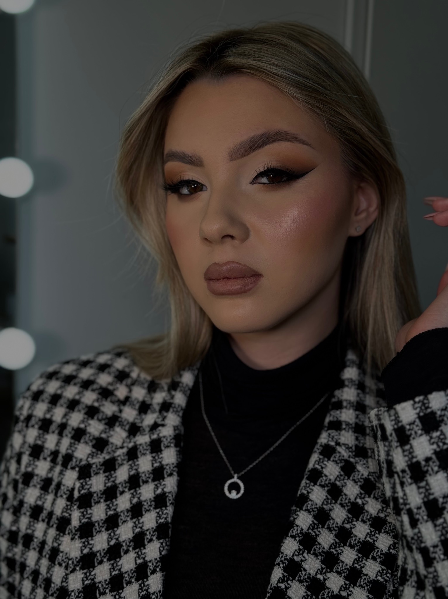 Portret kobiety z profesjonalnym, wieczorowym makijażem oczu z wyraźną kreską, podkreślonymi kośćmi policzkowymi i beżowymi ustami, ubrana w czarny golf i marynarkę w pepitkę, z delikatnym srebrnym...