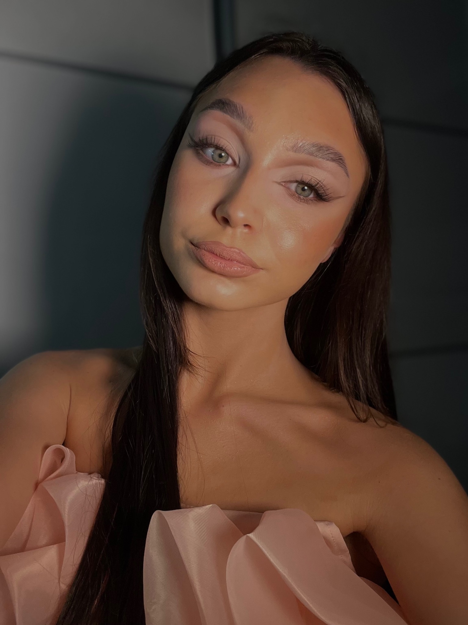 Portret kobiety z delikatnym, naturalnym upiększeniem, podkreślone oczy z subtelną kreską, jasnoróżowe usta, długie, ciemne włosy i bladoróżowa suknia z drapowaniem.