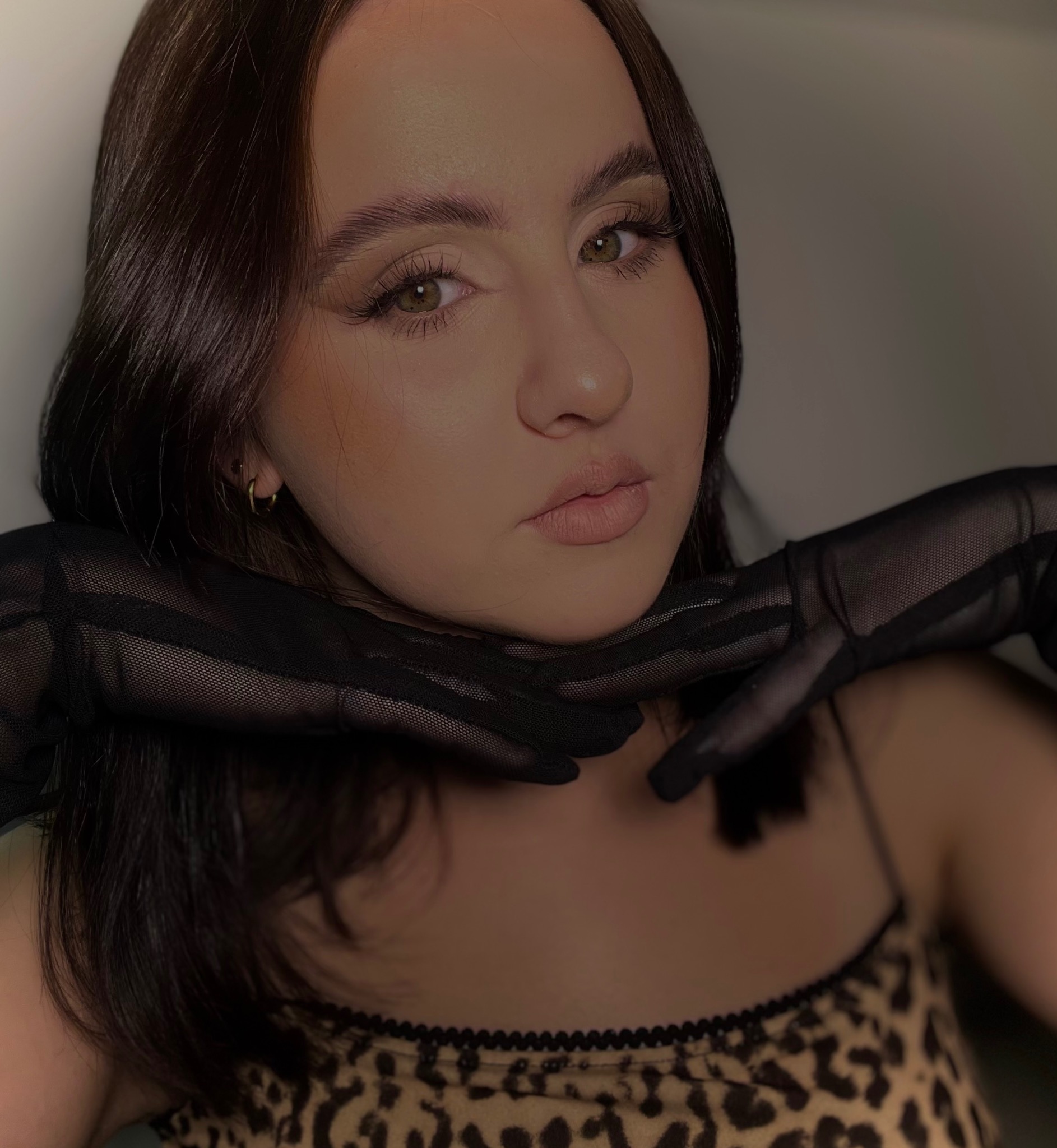 Portret kobiety w Warszawie z delikatnym makijażem, brązowe włosy, usta w odcieniu nude, dłonie w czarnych, siateczkowych rękawiczkach pod brodą, bluzka w panterkę.