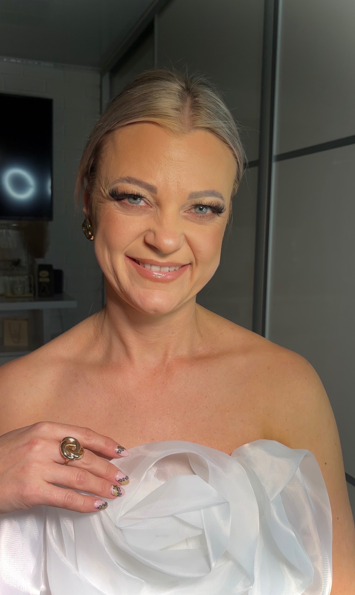 Uśmiechnięta blondynka z niebieskimi oczami, w delikatnym, błyszczącym makijażu ust i wydłużonych rzęsach, pozuje z dużą złotą biżuterią na dłoni i w uszach, ubrana w białą, zwiewną tkaninę.