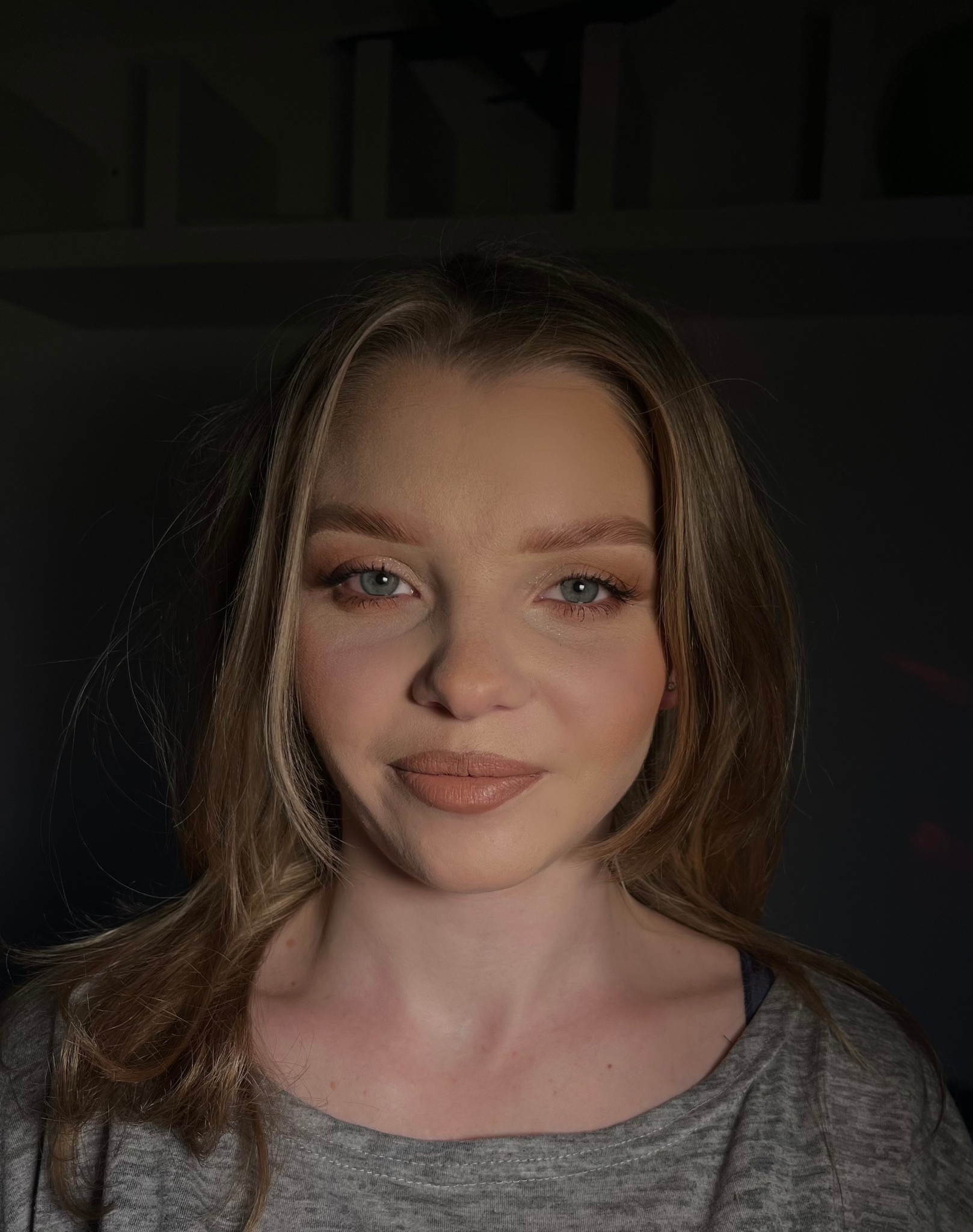 Portret kobiety z delikatnym, naturalnym makijażem w odcieniach nude, z podkreślonymi oczami i ustami, w ciemnym otoczeniu.