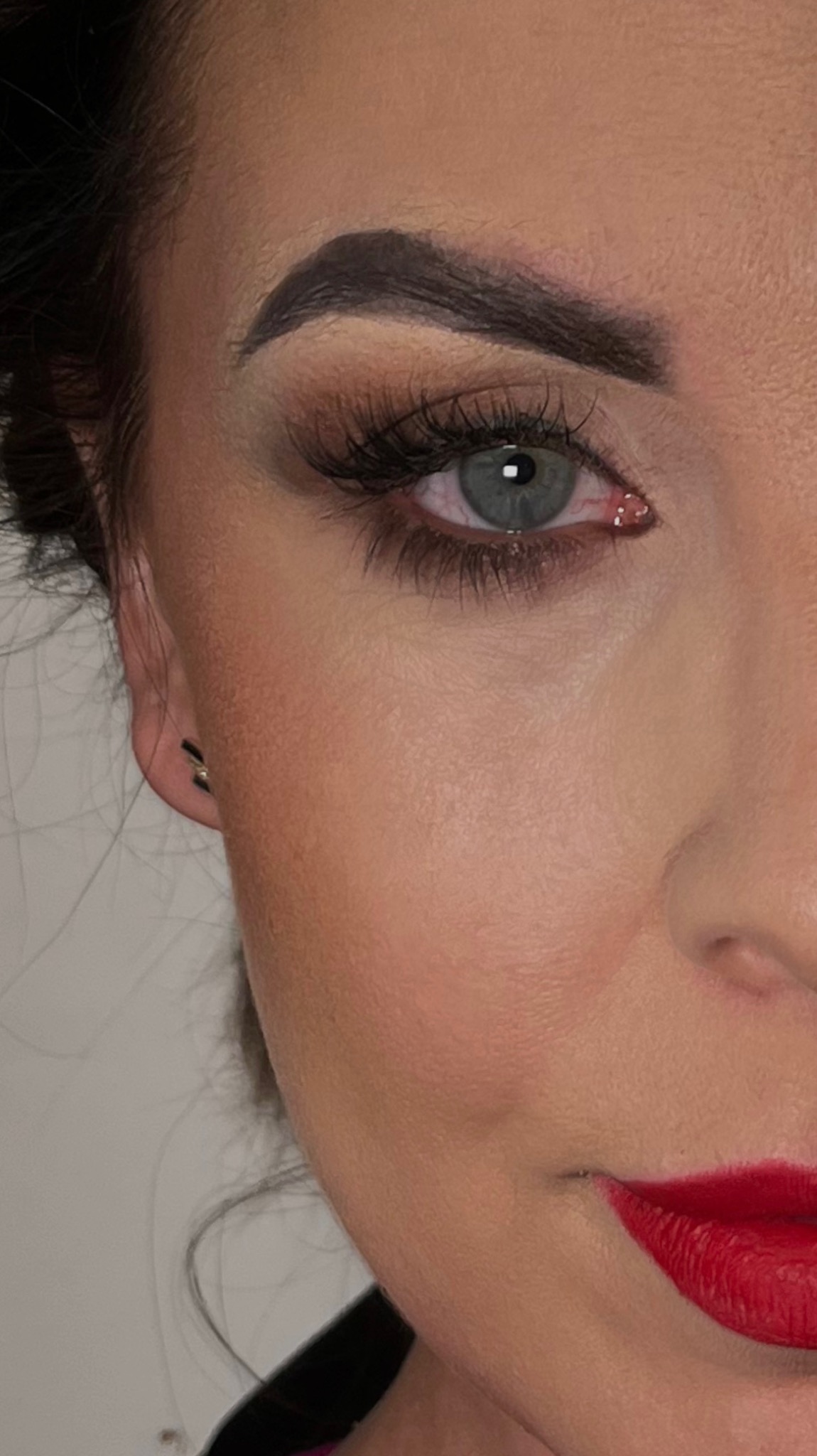 Fragment twarzy kobiety z intensywnym czerwonym kolorem ust, mocno podkreślonym okiem z długimi rzęsami i widocznym delikatnym cieniem, oraz precyzyjnie wymodelowanymi brwiami. W uchu widoczny mały...
