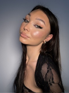 Portret młodej kobiety z wyrazistym makijażem oczu typu kocie oko, podkreślonymi brwiami i błyszczącymi ustami. Uwagę zwracają duże, złote kolczyki i czarna, koronkowa bluzka. Tło jest szare...