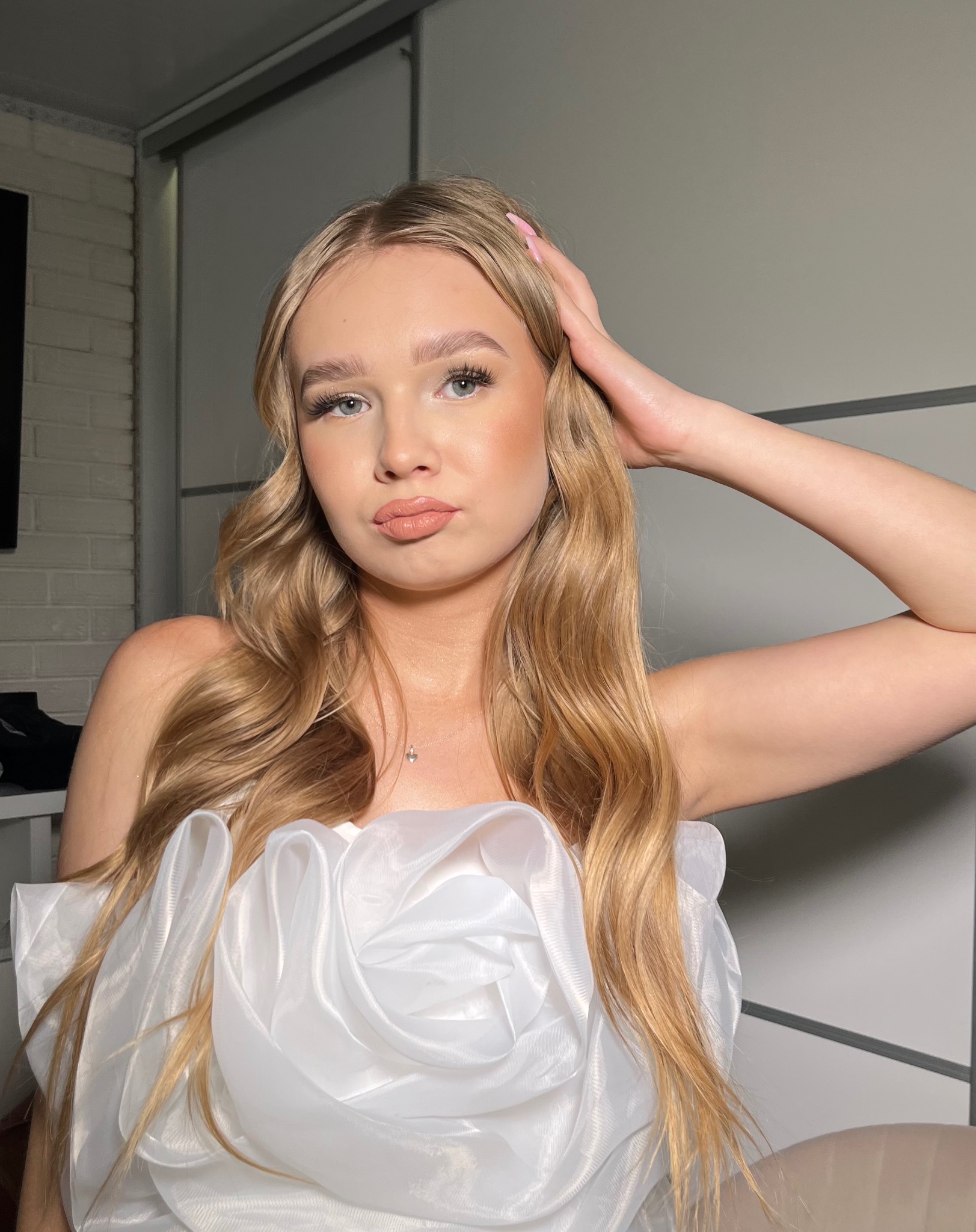 Portret młodej kobiety z blond włosami, delikatnym makijażem i białym, ozdobnym topem z organzy, pozującej na jasnym tle.