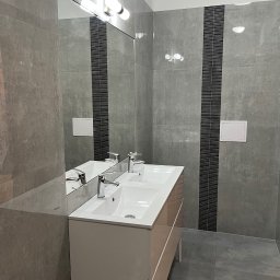 Usługi budowlane - Nowoczesna łazienka z podwójną umywalką, szare płytki imitujące beton, czarny dekoracyjny pas płytek, duże lustro z oświetleniem punktowym i biała zabudowa pod umywalką na metalowych nóżkach.