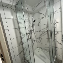 Usługi budowlane - Zmontowana kabina prysznicowa z chromowanymi okuciami, osadzona w narożniku łazienki wyłożonej białymi płytkami z szarym marmurkowym wzorem. Widoczny brodzik i słuchawka prysznicowa.