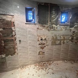 Usługi budowlane - Demontaż płytek w łazience z widocznymi fragmentami ścian i gruzem na podłodze. Dwa okna z widokiem na zewnątrz. Etap przygotowawczy do remontu.