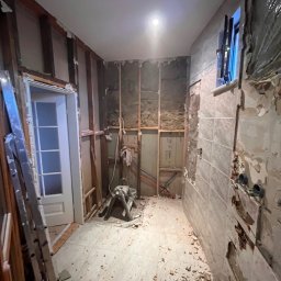 Usługi budowlane - Demontaż płytek w łazience, widoczne odsłonięte ściany z drewnianą konstrukcją, gruz na podłodze, drabina oparta o ścianę. Etap przygotowawczy do remontu.