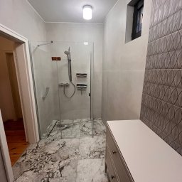 Usługi budowlane - Nowoczesna łazienka po remoncie: kabina prysznicowa ze szklanymi drzwiami, marmurowe płytki podłogowe i szafka z szufladami. Dekoracyjna ściana z płytek.