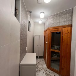 Usługi budowlane - Nowoczesna łazienka z drewnianą sauną, marmurową podłogą i szafką w odcieniach szarości. Geometryczne płytki na ścianie dodają elegancji. Oświetlenie sufitowe.