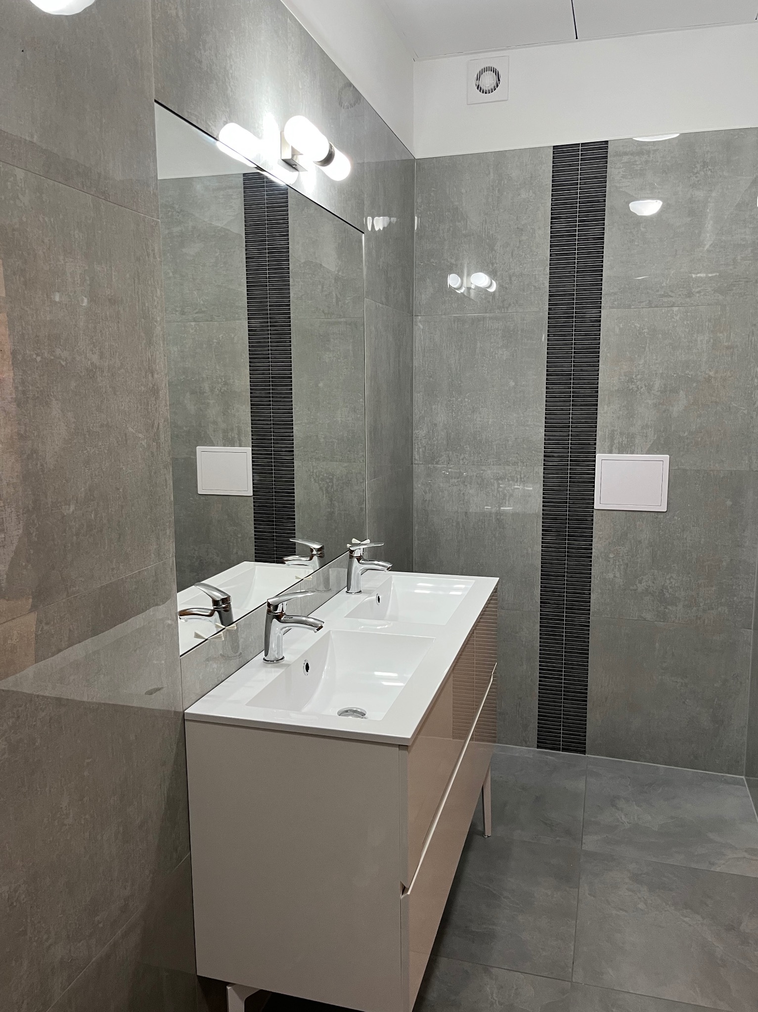 Nowoczesna łazienka z podwójną umywalką, szare płytki imitujące beton, czarny dekoracyjny pas płytek, duże lustro z oświetleniem punktowym i biała zabudowa pod umywalką na metalowych nóżkach.