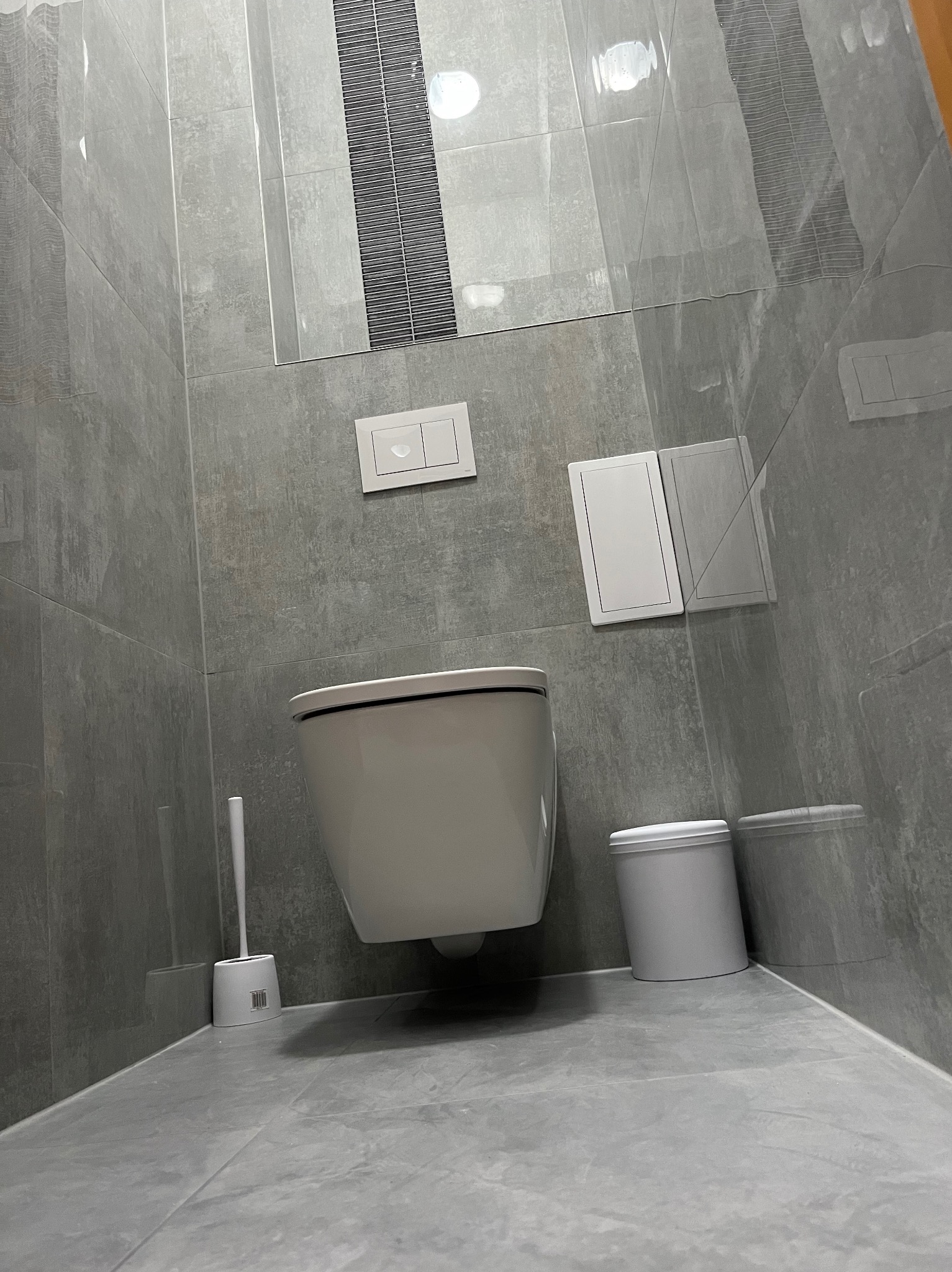 Nowoczesna łazienka z szarymi płytkami imitującymi kamień, podwieszana toaleta z białym przyciskiem spłukiwania, biały pojemnik na szczotkę WC i kratka wentylacyjna w zabudowie.