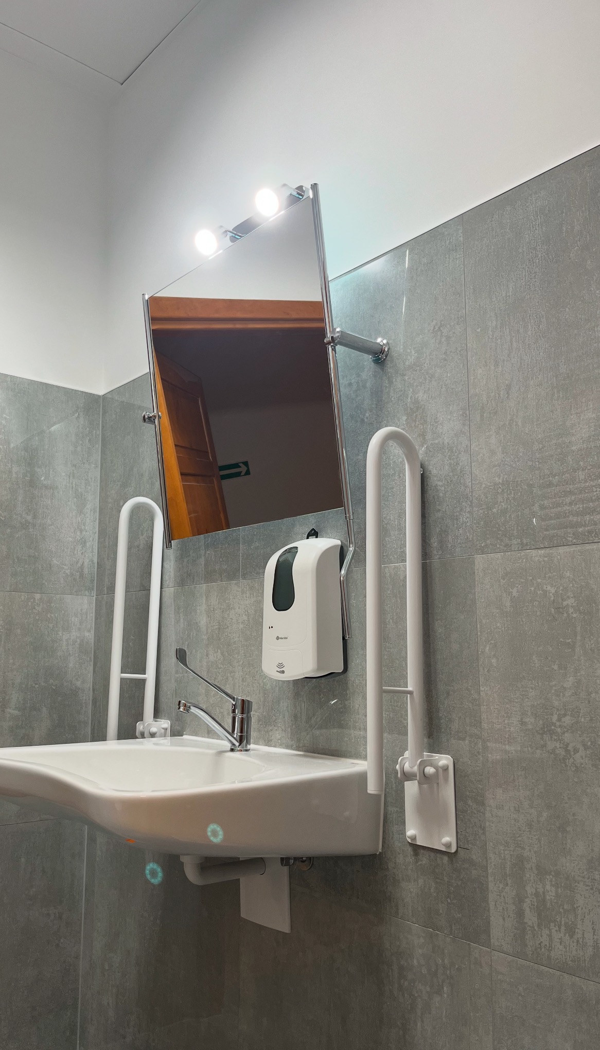 Umywalka dla niepełnosprawnych z uchwytami, dozownikiem mydła i lustrem z oświetleniem, na tle szarych płytek ceramicznych, widoczny fragment drzwi w odbiciu lustra.