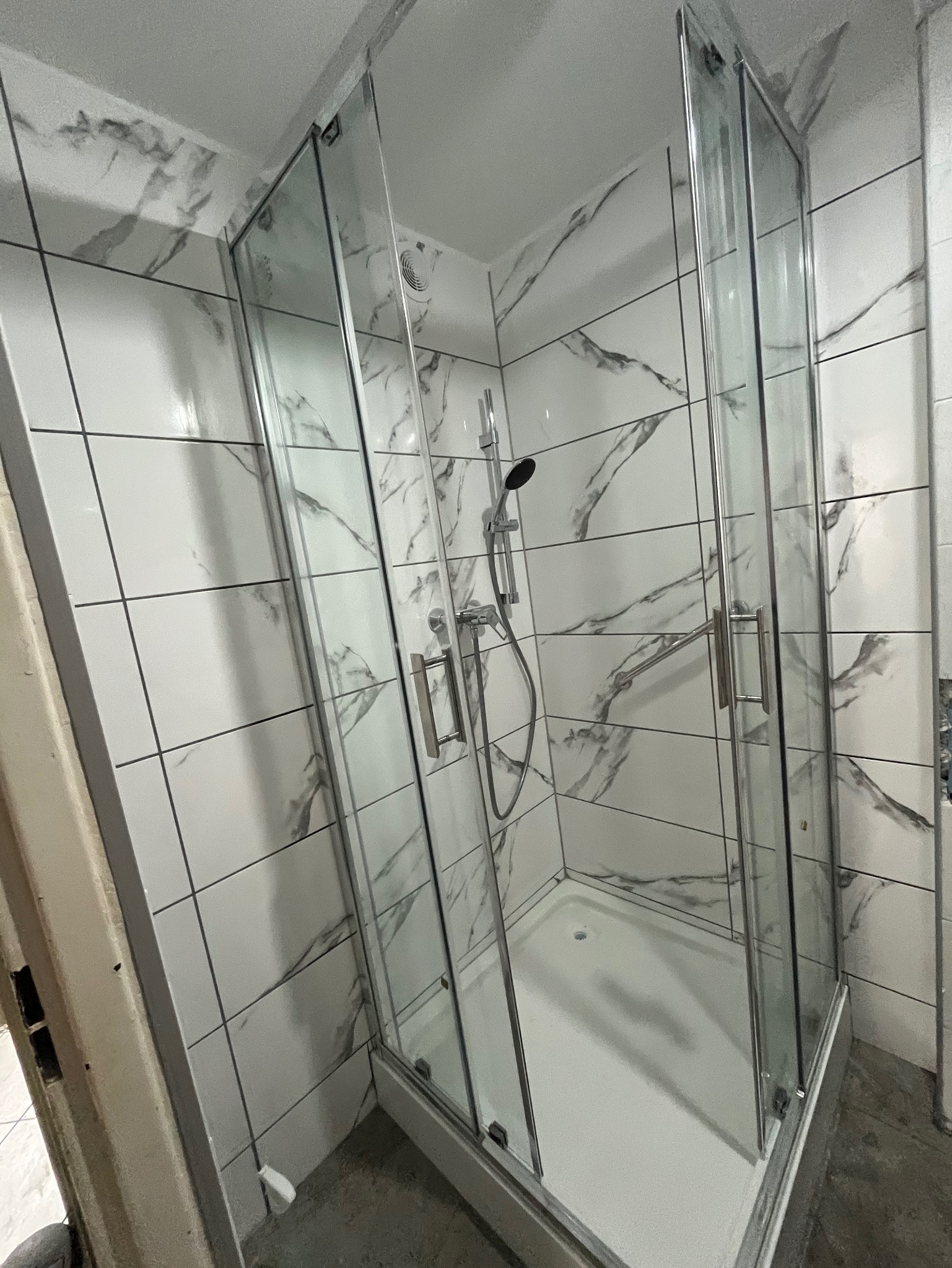 Zmontowana kabina prysznicowa z chromowanymi okuciami, osadzona w narożniku łazienki wyłożonej białymi płytkami z szarym marmurkowym wzorem. Widoczny brodzik i słuchawka prysznicowa.