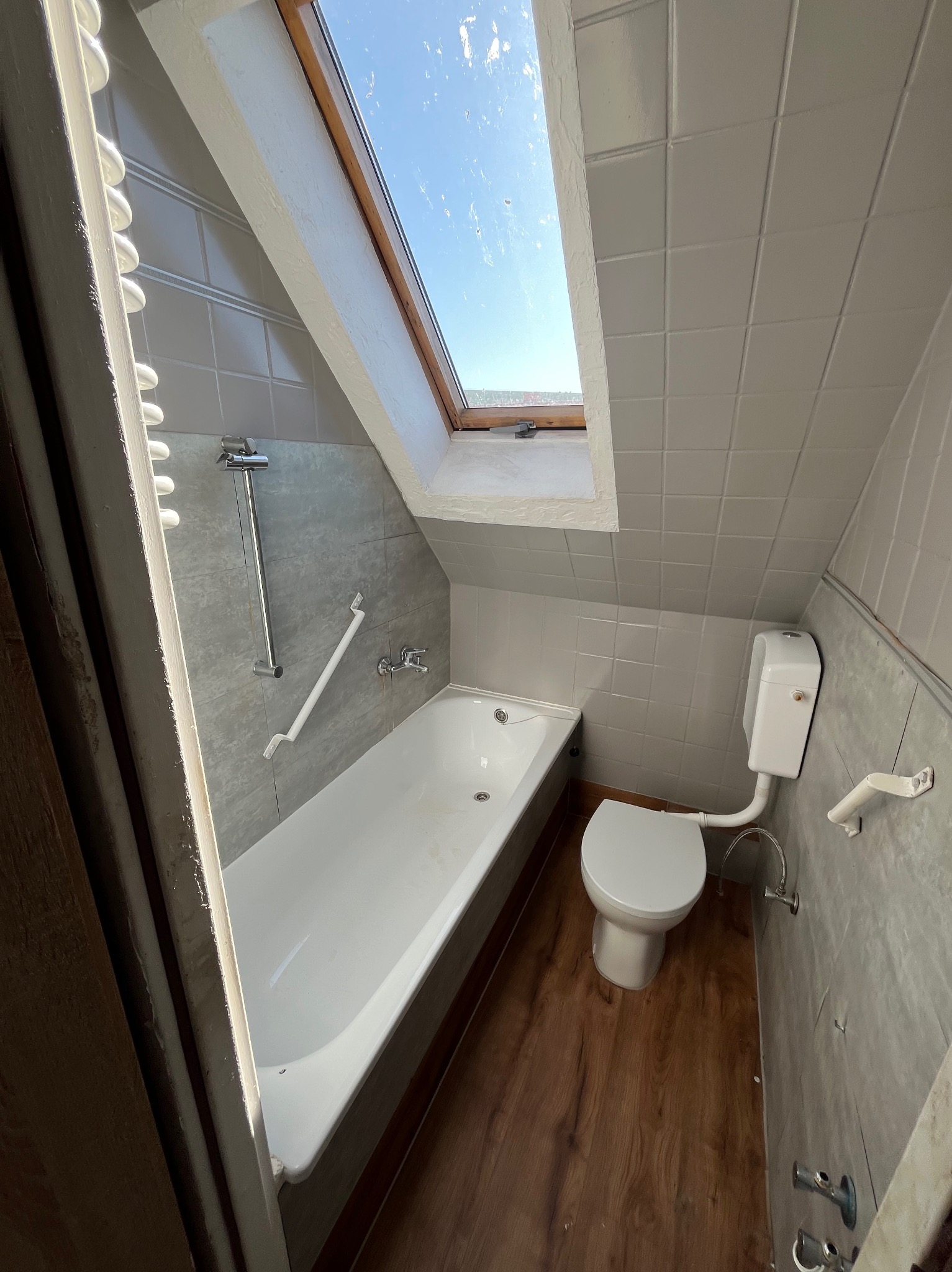 Szara łazienka na poddaszu z oknem dachowym, białą wanną i toaletą. Uwagę zwraca drewnopodobna podłoga i płytki na ścianach w odcieniach szarości.