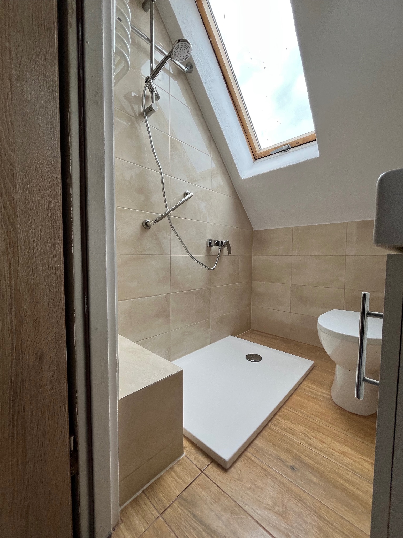 Jasna łazienka na poddaszu z kabiną prysznicową, beżowymi płytkami i oknem dachowym. Widok zza uchylonych, drewnianych drzwi.