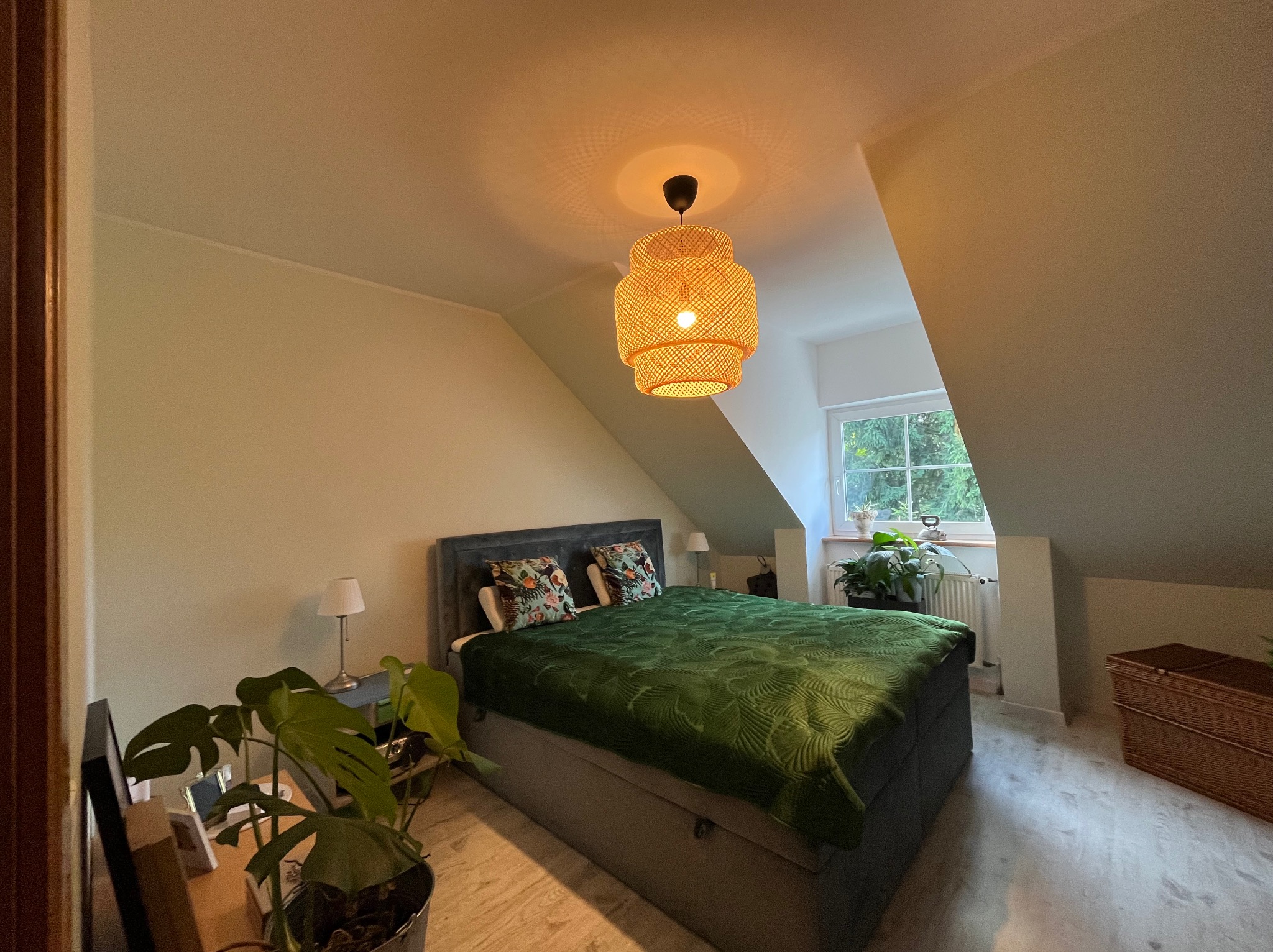 Sypialnia na poddaszu z łóżkiem, roślinami i plecioną lampą w stylu boho. Ściany w odcieniach beżu. Widoczne skosy i okno z widokiem na zieleń.