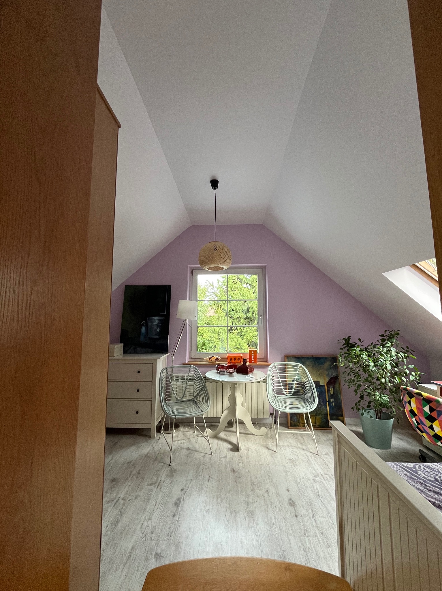 Jasne poddasze z fioletowymi ścianami, oknem z widokiem na zieleń, okrągłym stołem z krzesłami i drewnianą podłogą. Uwagę przykuwa lampa wisząca nad stołem.