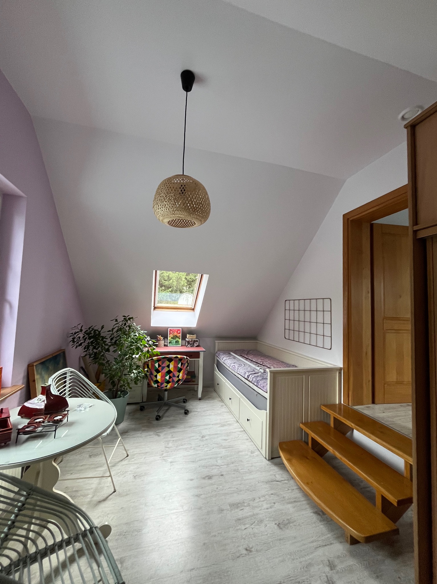 Jasne poddasze ze skosem i oknem dachowym. Widoczny stół z krzesłami, biurko z kolorowym krzesłem, łóżko z szufladami i drewniane schody.