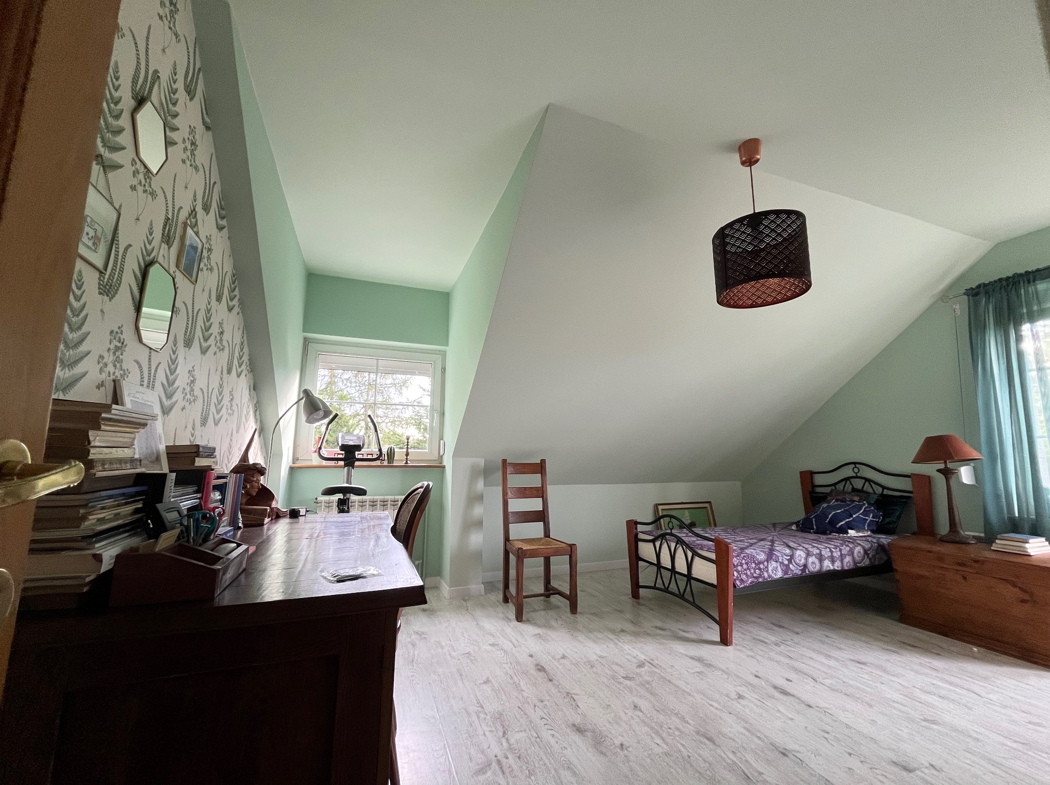 Pokój na poddaszu z biurkiem, łóżkiem i zielonymi ścianami. Tapeta w liście, drewniana podłoga, skośny sufit i okno z widokiem na zewnątrz.