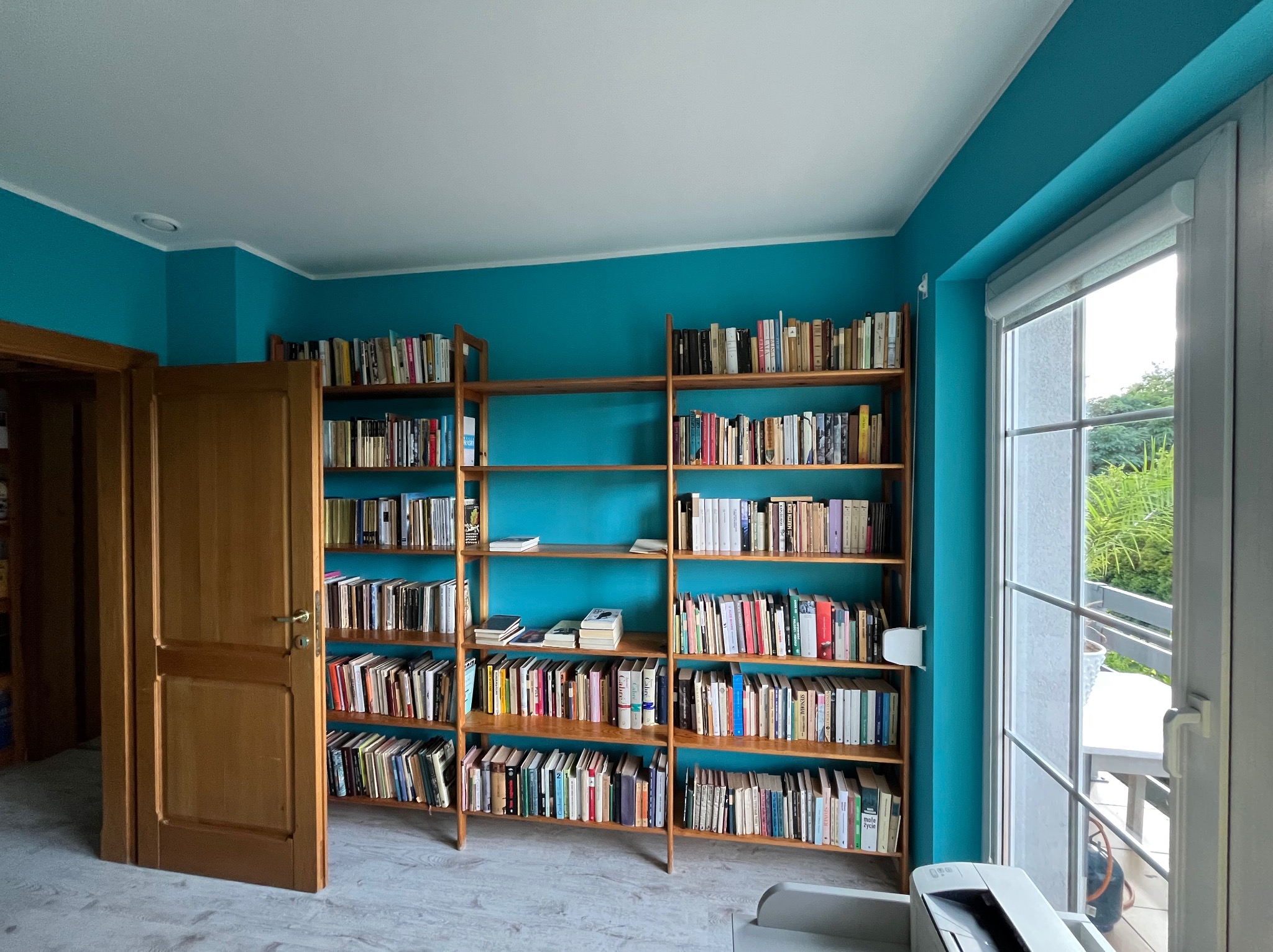 Turkusowe wnętrze z drewnianym regałem pełnym książek. Widoczne drzwi i okno balkonowe z roletą. Podłoga w jasnym odcieniu, drukarka w rogu.