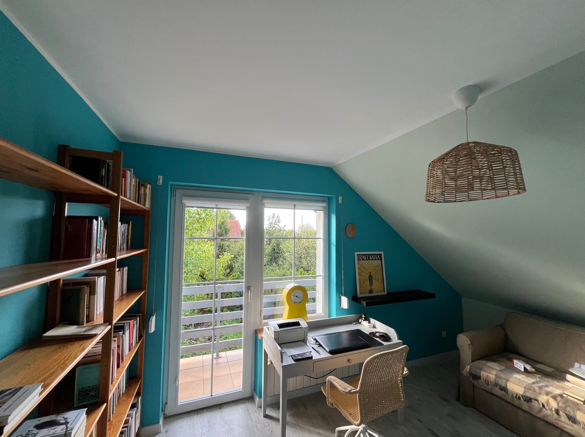 Turkusowe ściany i biały sufit w pokoju na poddaszu z biurkiem, regałem z książkami i wyjściem na balkon. Wnętrze po remoncie, jasne, z elementami dekoracyjnymi.