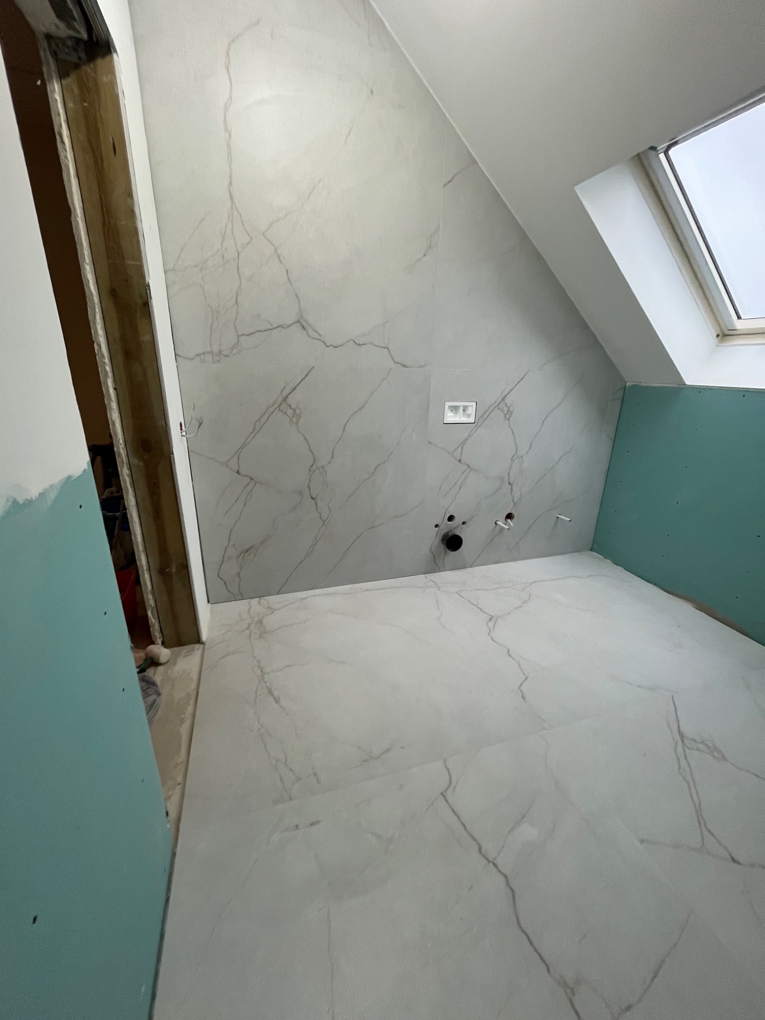 Łazienka na poddaszu w trakcie remontu: białe płytki imitujące marmur na podłodze i ścianach, widoczne instalacje, okno dachowe i płyty gipsowo-kartonowe.