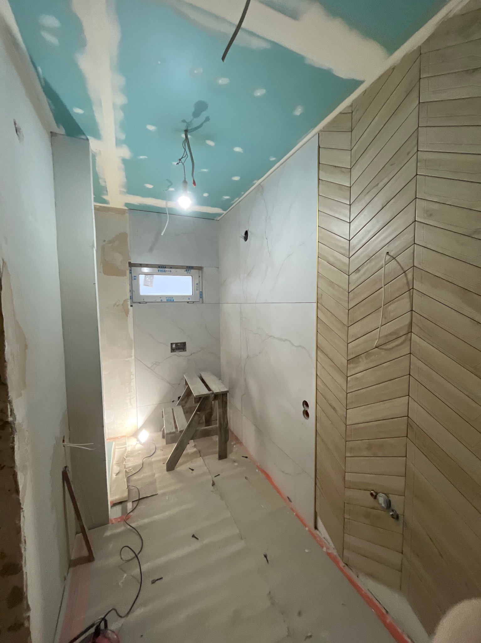Wnętrze łazienki w trakcie wykańczania: położone płytki imitujące marmur i drewno w jodełkę, sufit z płyt karton-gips, drabina, okno. Widoczne przewody i elementy instalacji.