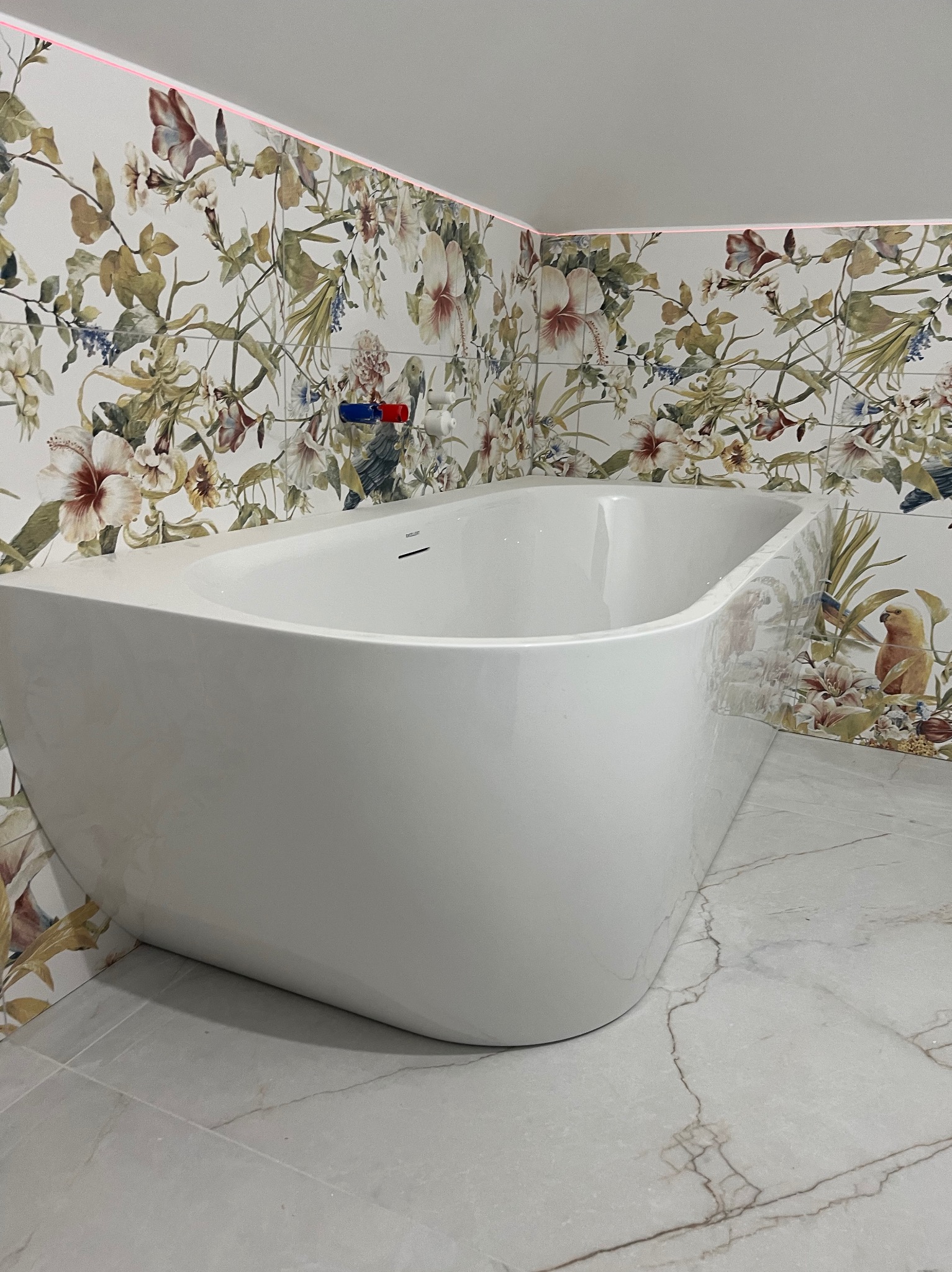 Nowoczesna, biała wanna wolnostojąca w łazience z kwiatowymi płytkami. Marmurowa podłoga i subtelne oświetlenie LED podkreślają elegancję wykończenia.