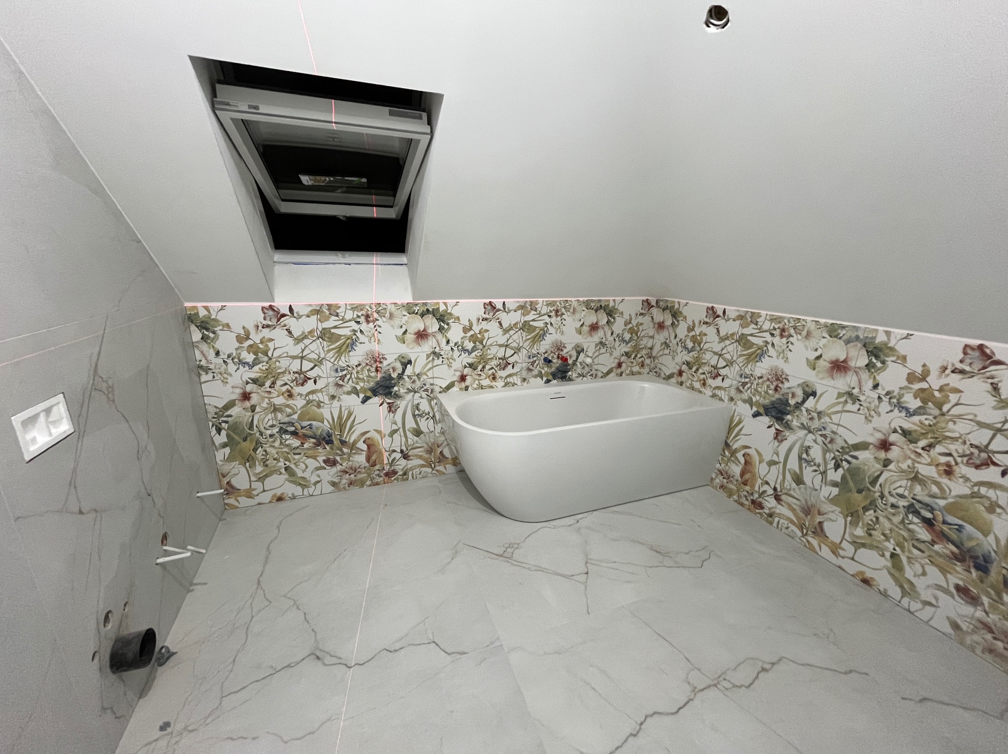 Biała wanna wolnostojąca w łazience na poddaszu z oknem dachowym. Ściany wyłożone płytkami z motywem kwiatowym i marmurową podłogą. Widoczne instalacje.