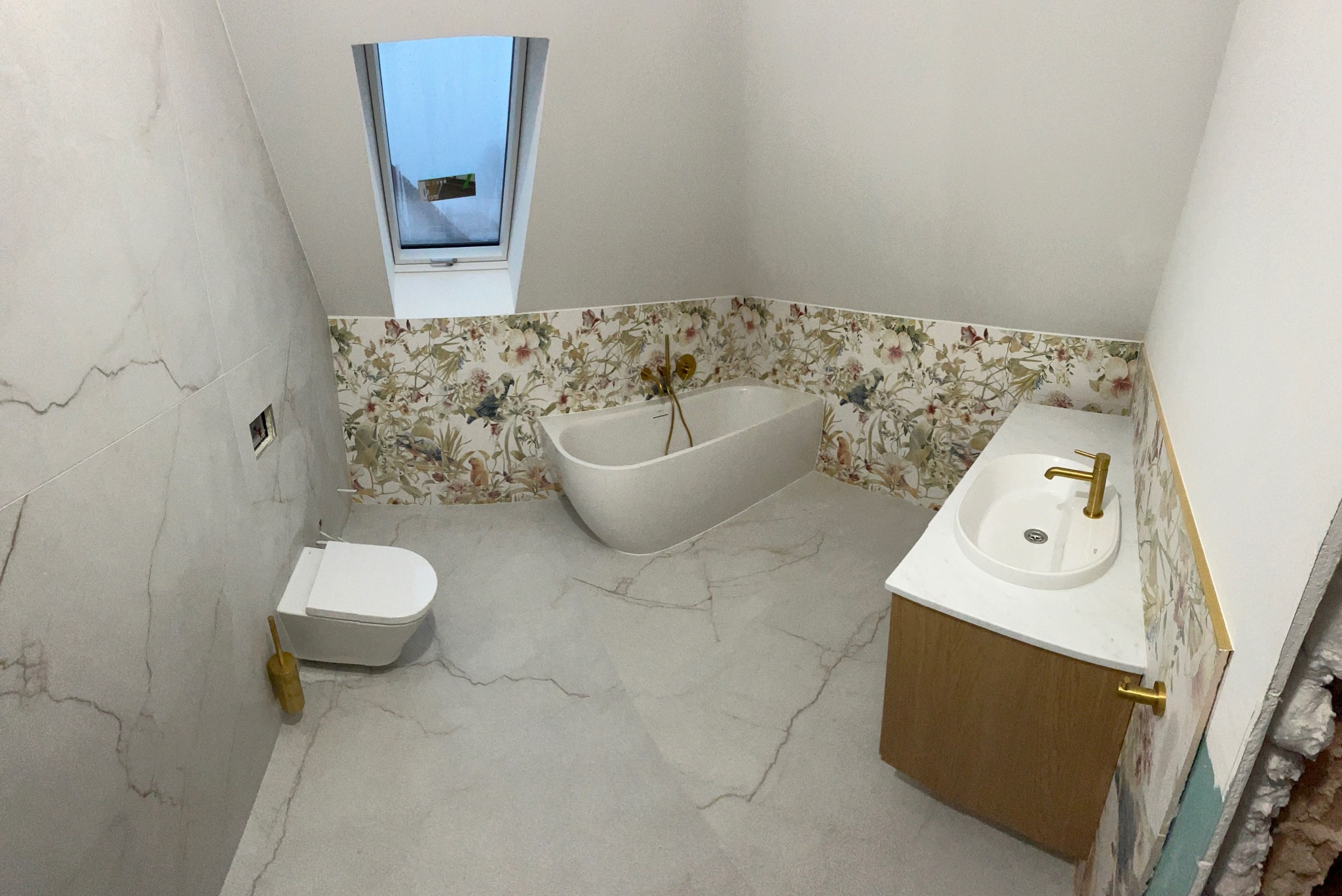 Wykończona łazienka na poddaszu z wanną, umywalką z szafką i toaletą. Ściany wyłożone płytkami imitującymi marmur i płytkami z motywem kwiatowym.