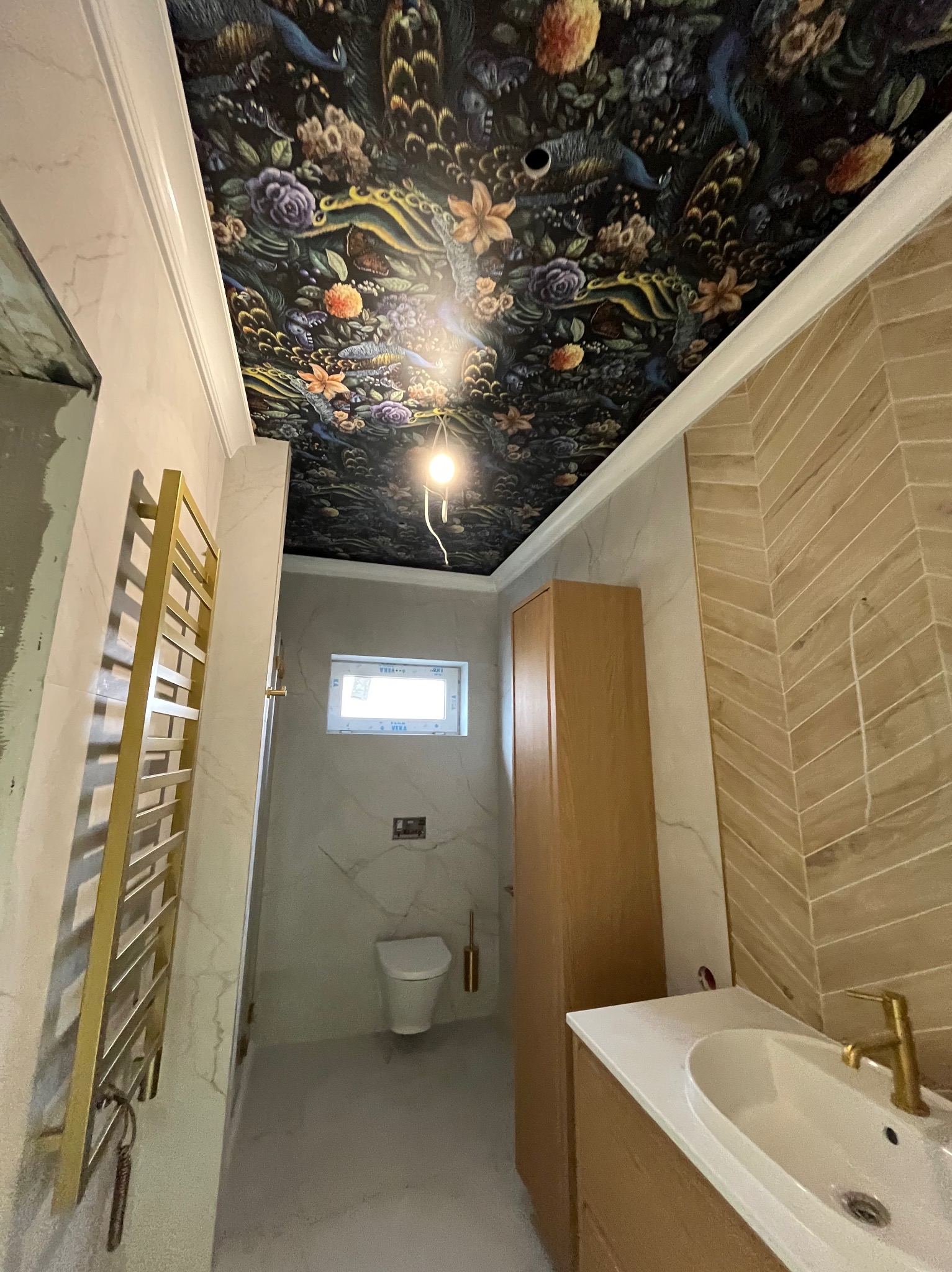 Wykończona łazienka z wzorzystym sufitem, drewnianymi szafkami i grzejnikiem w kolorze złota. Na ścianach płytki imitujące marmur i drewno ułożone w jodełkę.