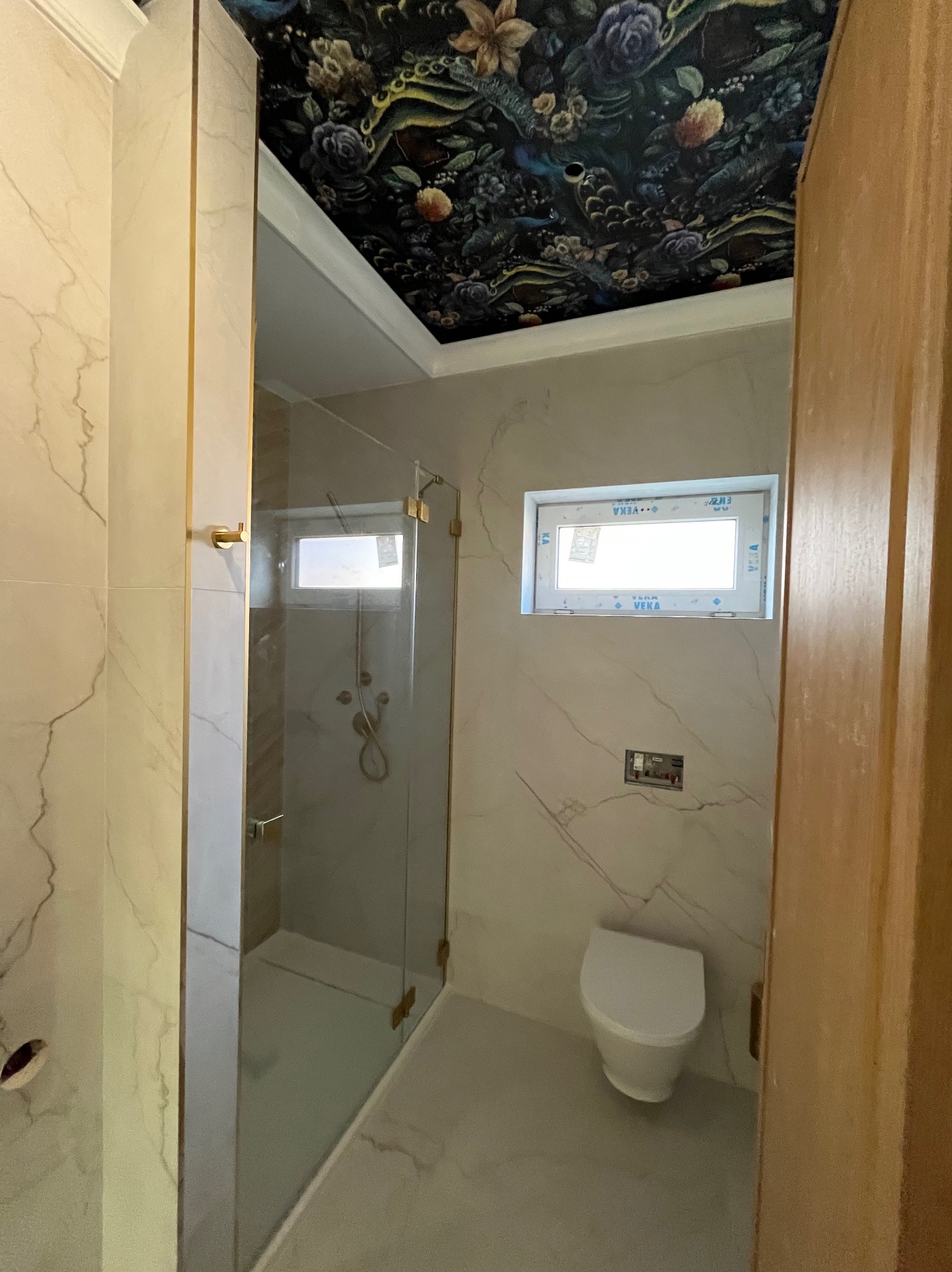 Wykończona łazienka z kabiną prysznicową, toaletą i oknem. Ściany wyłożone płytkami imitującymi marmur, sufit z motywem kwiatowym. Złote akcenty w kabinie.
