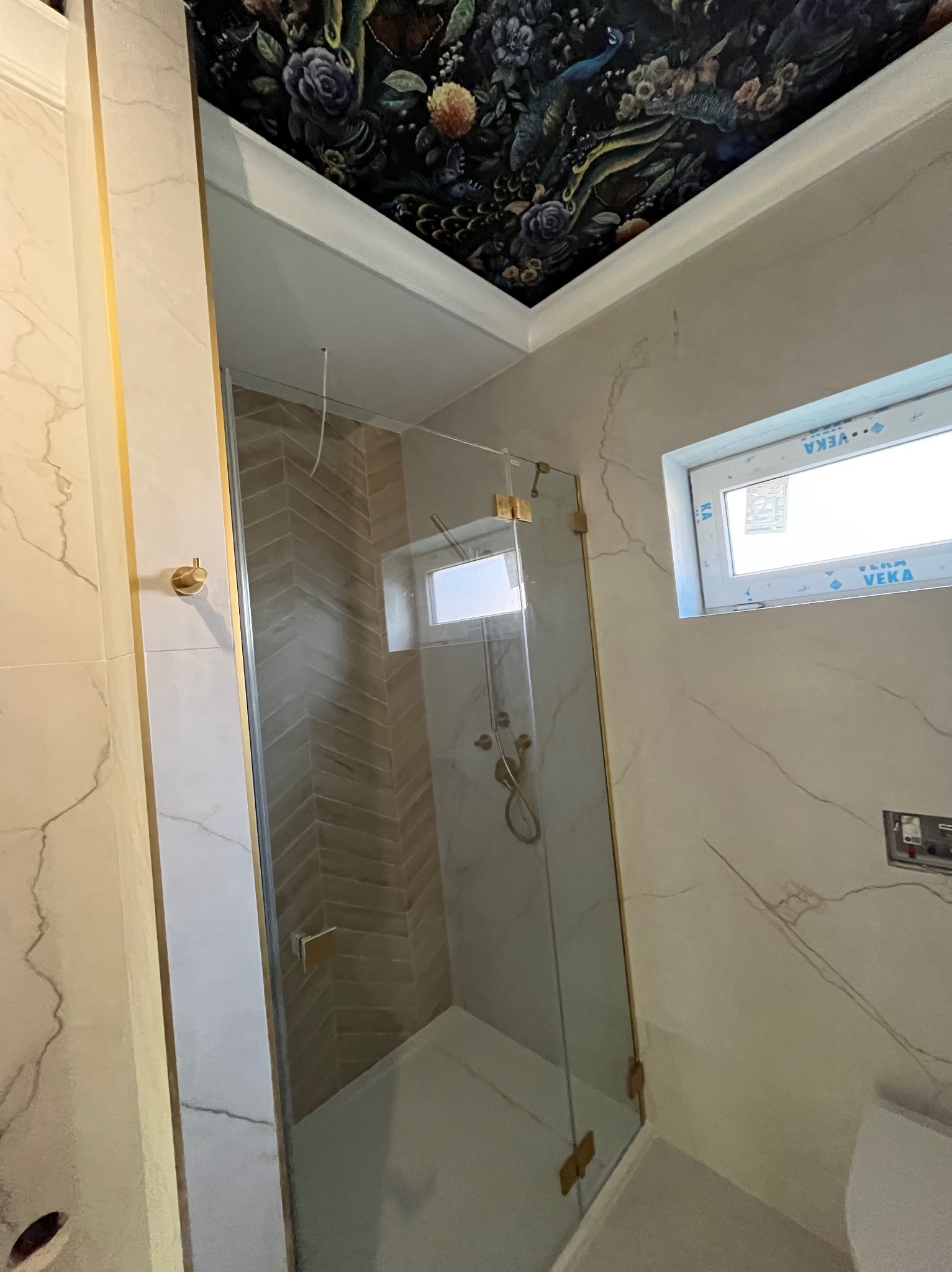 Nowoczesna kabina prysznicowa z złotymi detalami i wzorzystym sufitem w łazience wykończonej marmurowymi płytkami. Widoczne okno i armatura.