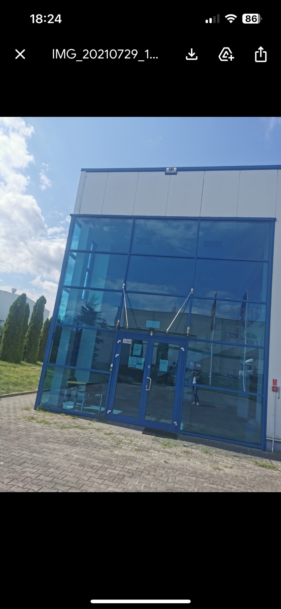 Szklana fasada budynku biurowego z niebieskimi ramami okiennymi, odbicie osoby w szybie, widoczna kostka brukowa przed wejściem.