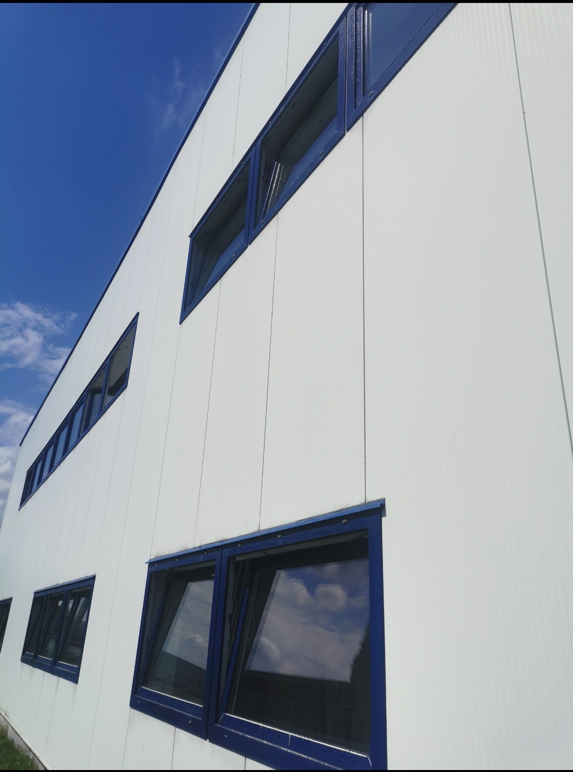 Biała elewacja budynku przemysłowego z rzędem okien w granatowych ramach, odbicie chmur na szybach, widok z dołu pod kątem.
