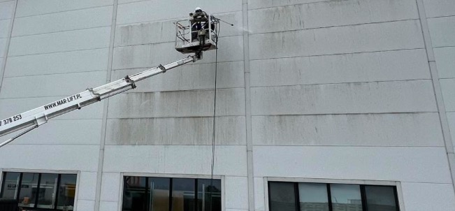 Pracownik w podnośniku koszowym czyści elewację budynku przemysłowego za pomocą myjki ciśnieniowej, widoczne zacieki i brud na ścianie przed czyszczeniem.