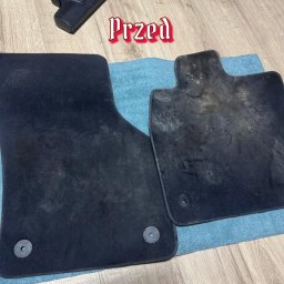 Czysta tapicera - Dwa czarne dywaniki samochodowe na niebieskiej ścierce, jeden czysty, drugi brudny, z napisem 'Przed' i odkurzaczem w tle. Ukazują efekt przed czyszczeniem.