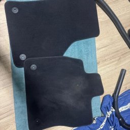 Czysta tapicera - Czarne, wyczyszczone dywaniki samochodowe leżące na niebieskim ręczniku, obok widoczny odkurzacz. Zdjęcie 'po' czyszczeniu, na drewnianej podłodze.