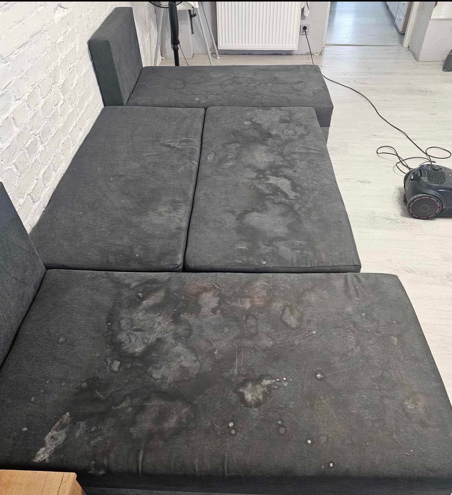 Szara, modułowa sofa z widocznymi plamami przed czyszczeniem, obok odkurzacz piorący. Widoczna ceglana ściana i jasna podłoga.