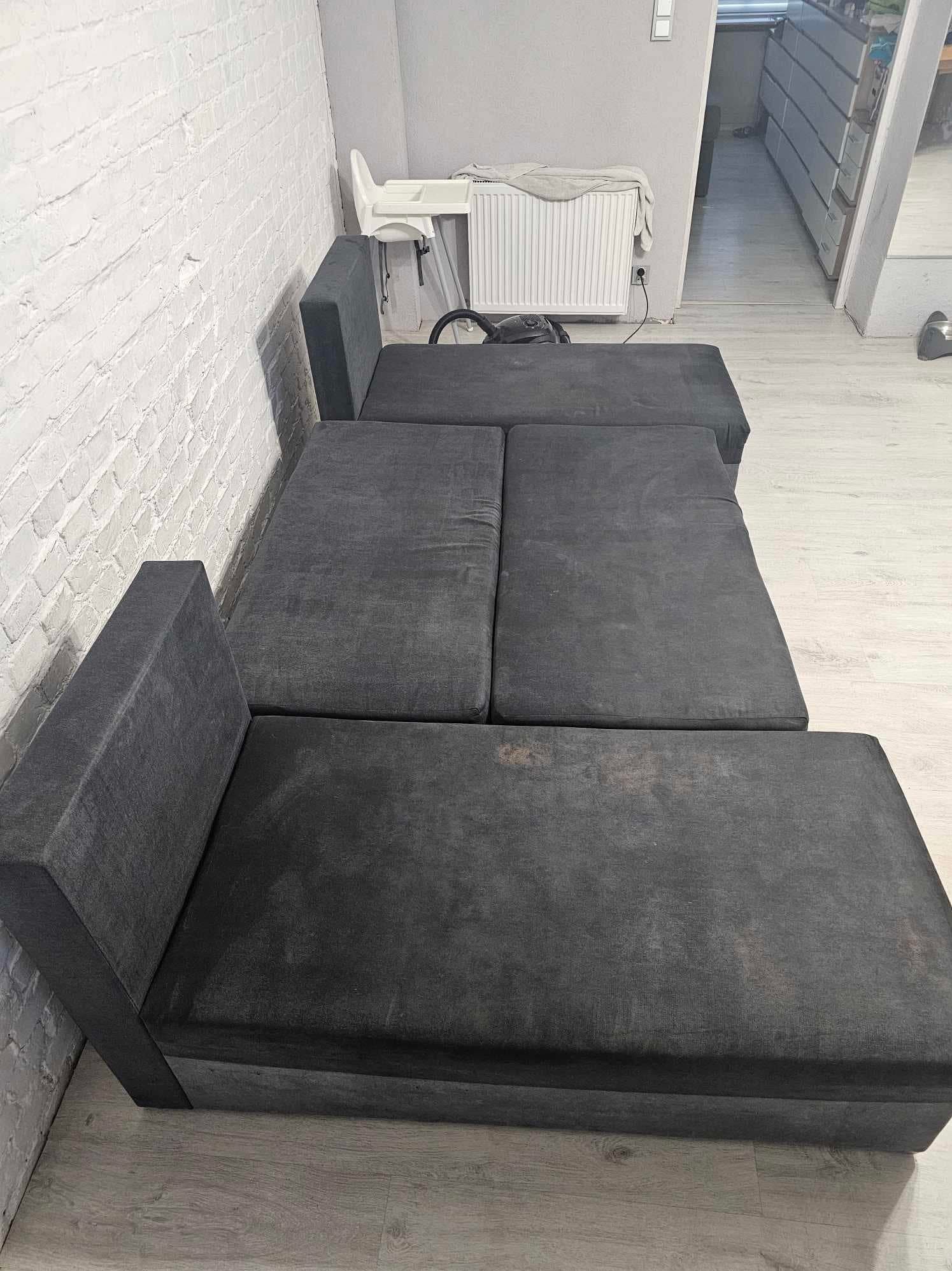 Rozłożona, szara kanapa modułowa z widocznymi plamami na tapicerce, ustawiona w rogu pokoju z białą ceglaną ścianą i jasną podłogą.