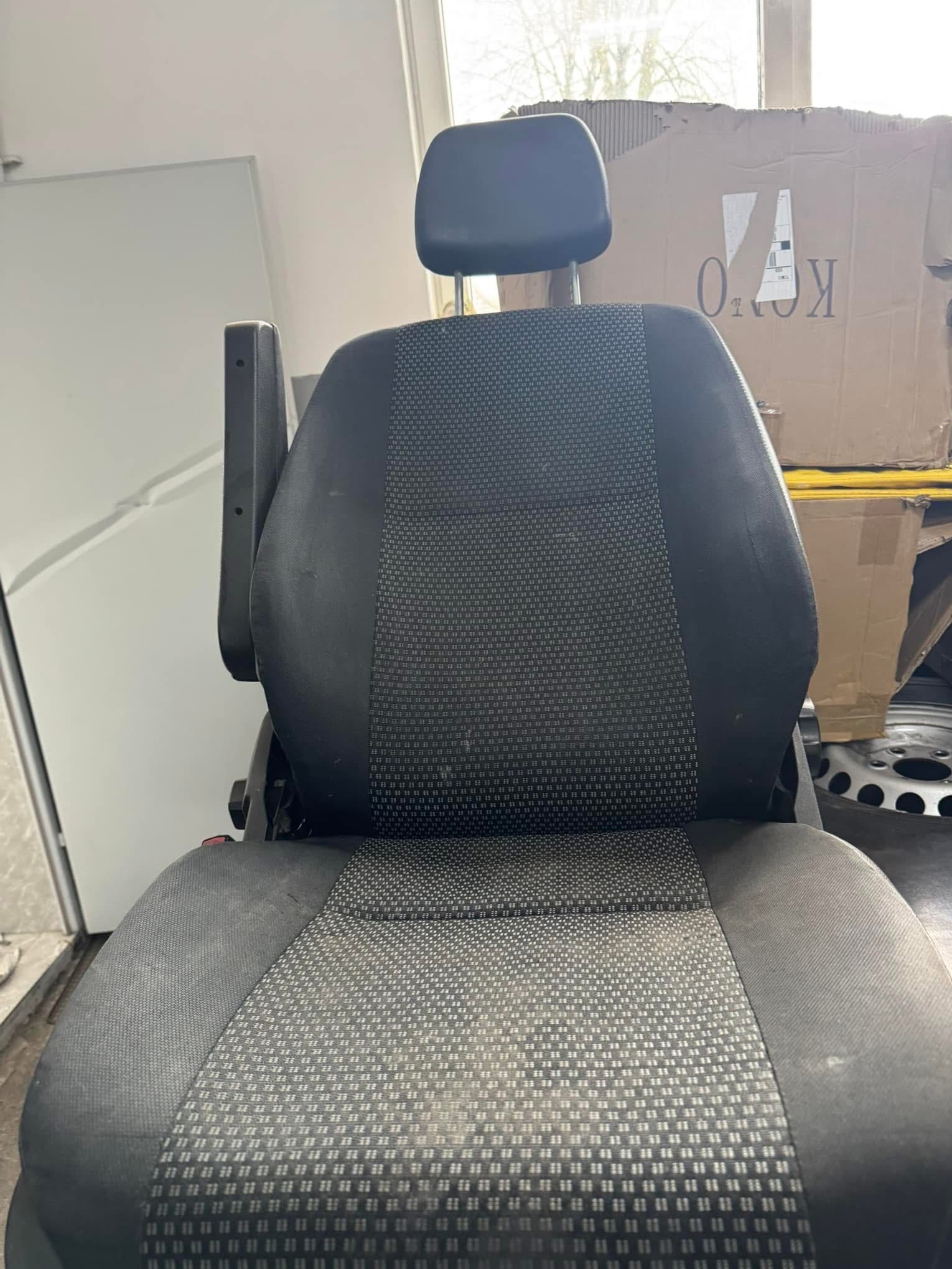 Brudne, szare siedzenie samochodowe z podłokietnikiem, widoczne plamy i zabrudzenia na tapicerce. W tle kartonowe pudła i fragment okna.