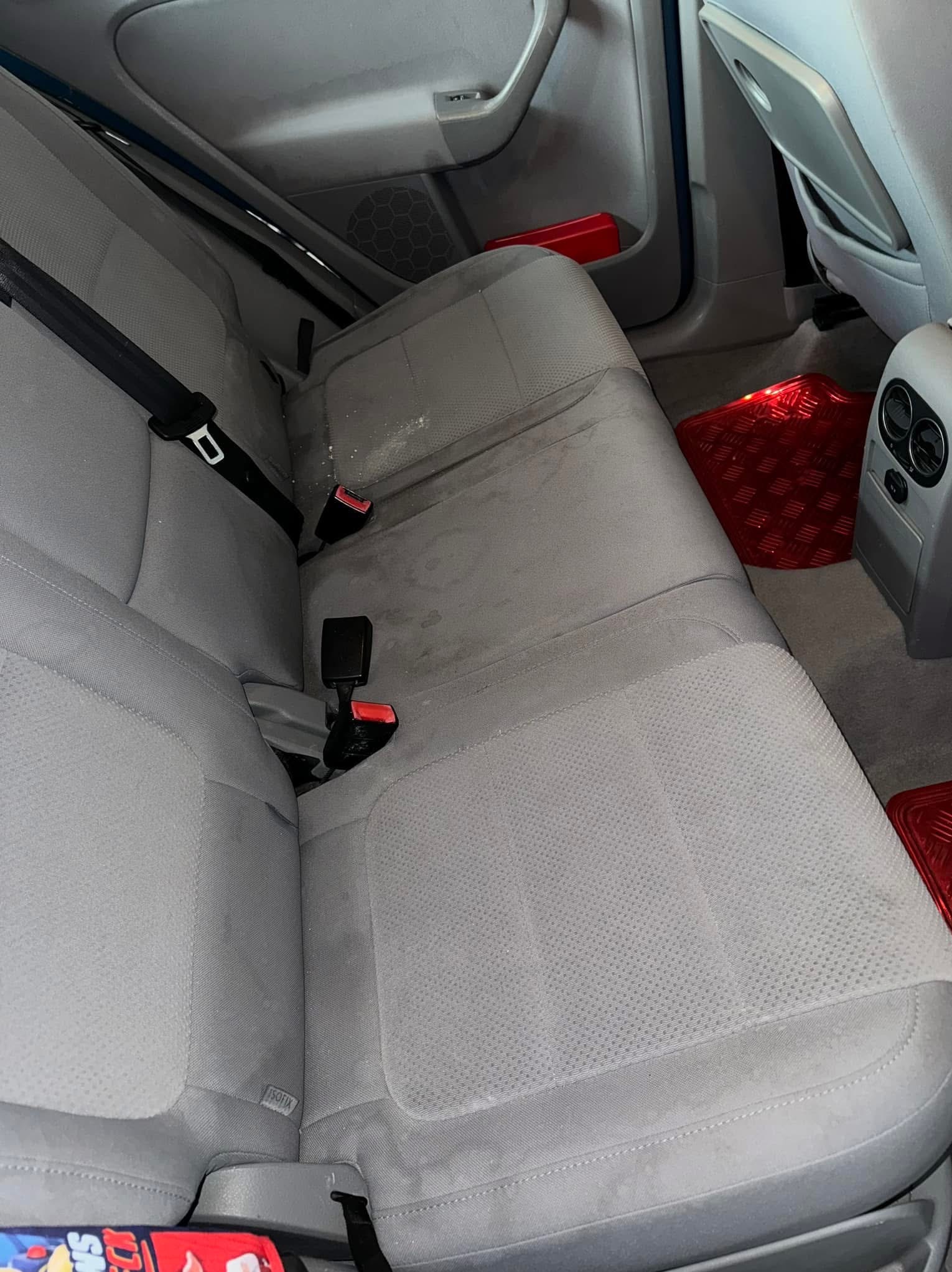 Szare, materiałowe siedzenie samochodowe z widocznymi plamami. Czerwone dywaniki pod nogami. Widoczne pasy bezpieczeństwa i elementy wnętrza pojazdu.