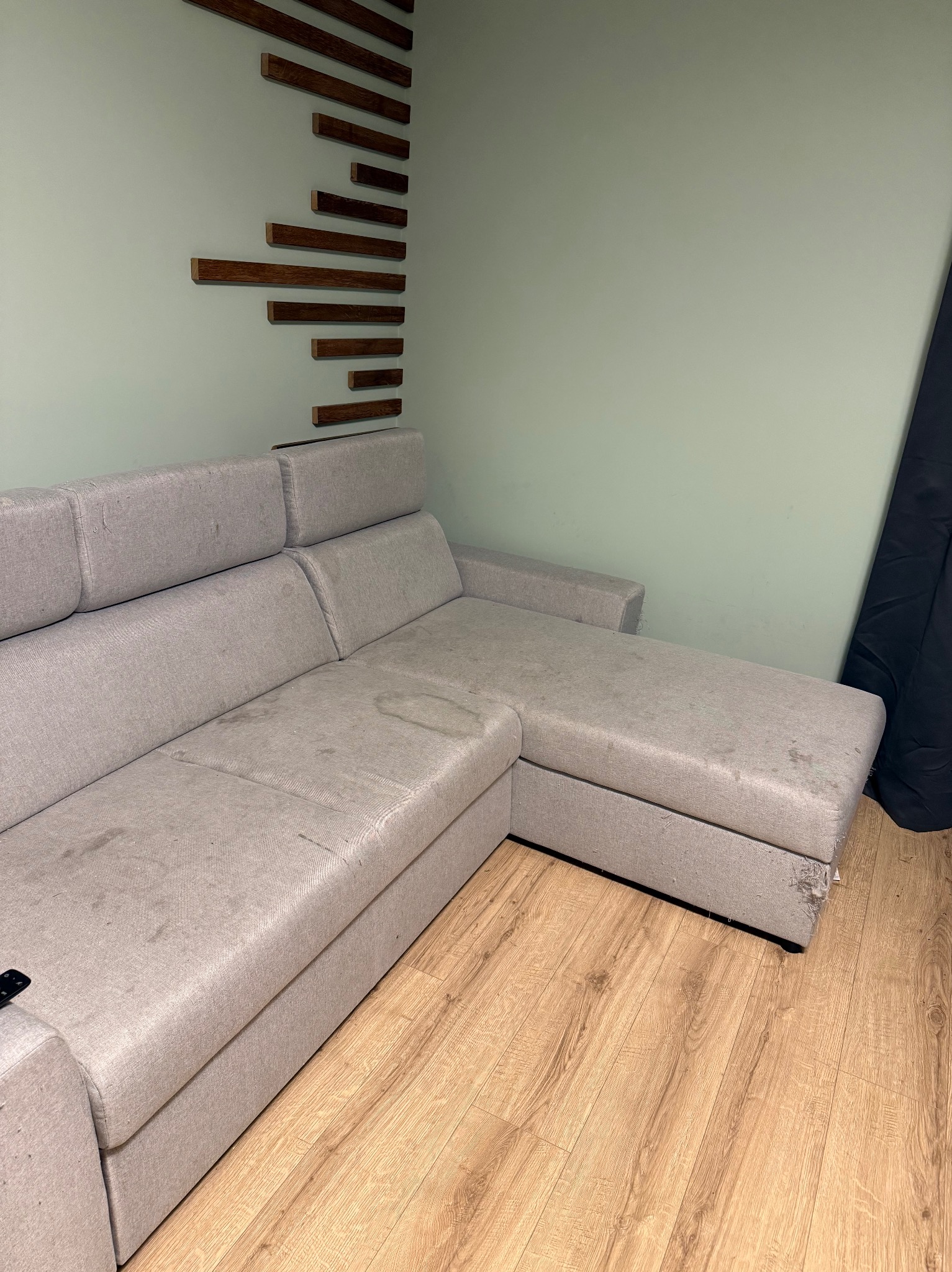 Szara, narożna kanapa z widocznymi plamami na tapicerce, stojąca na drewnianej podłodze. Na ścianie dekoracja z drewnianych listewek. Widoczne uszkodzenie tapicerki.