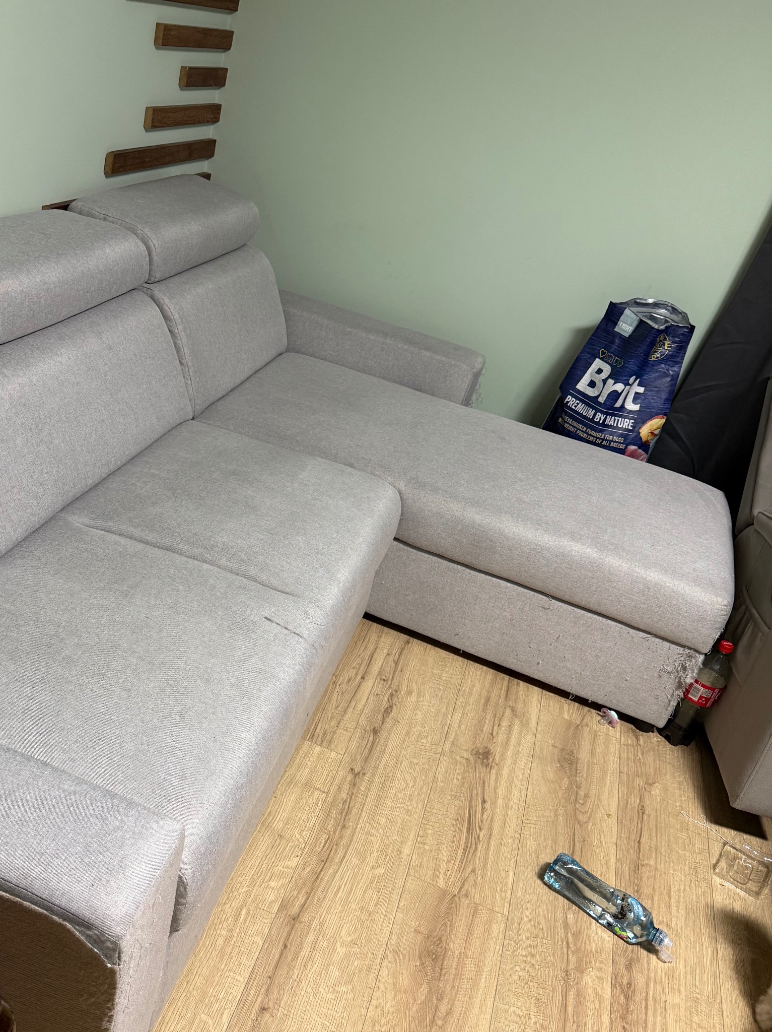 Szara, narożna kanapa z widocznymi śladami użytkowania, butelka wody i karma dla psa w tle. Widoczne zabrudzenia i zniszczenia tapicerki.