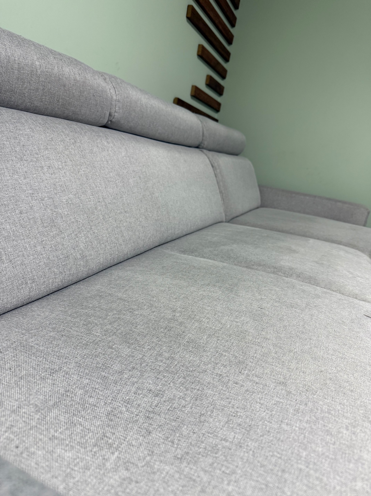 Szczegółowe ujęcie szarej, modułowej sofy z zagłówkami na tle pistacjowej ściany z drewnianą dekoracją. Widoczna faktura tkaniny i precyzyjne szwy.
