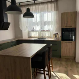 Nowoczesna kuchnia z wyspą, zielonymi szafkami i drewnianymi blatami. Nad wyspą wiszą dwie czarne lampy. Uwagę zwraca jasna podłoga i światło słoneczne.