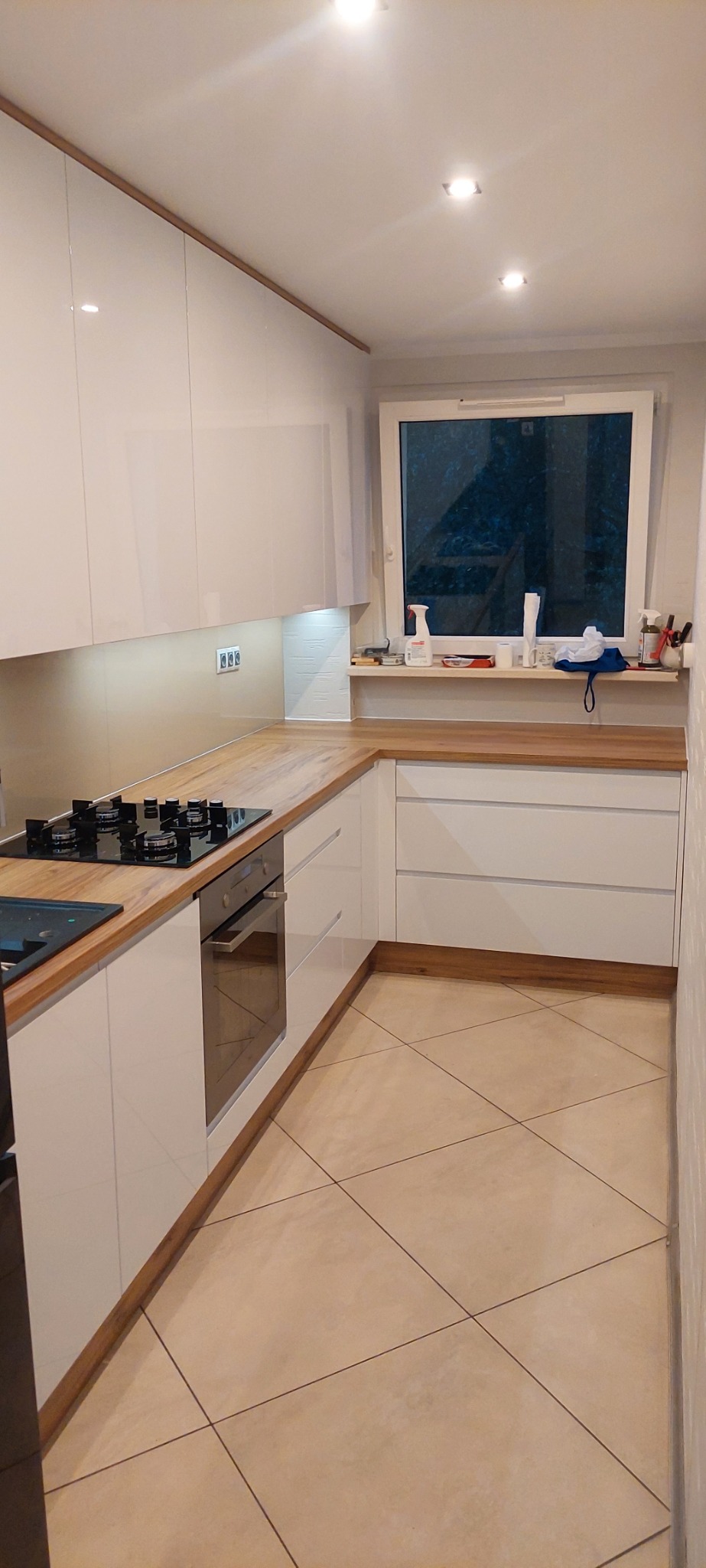 Nowoczesna kuchnia z białymi, lakierowanymi frontami i drewnianym blatem. Widoczna płyta grzewcza i piekarnik. Nad blatem okno z akcesoriami.