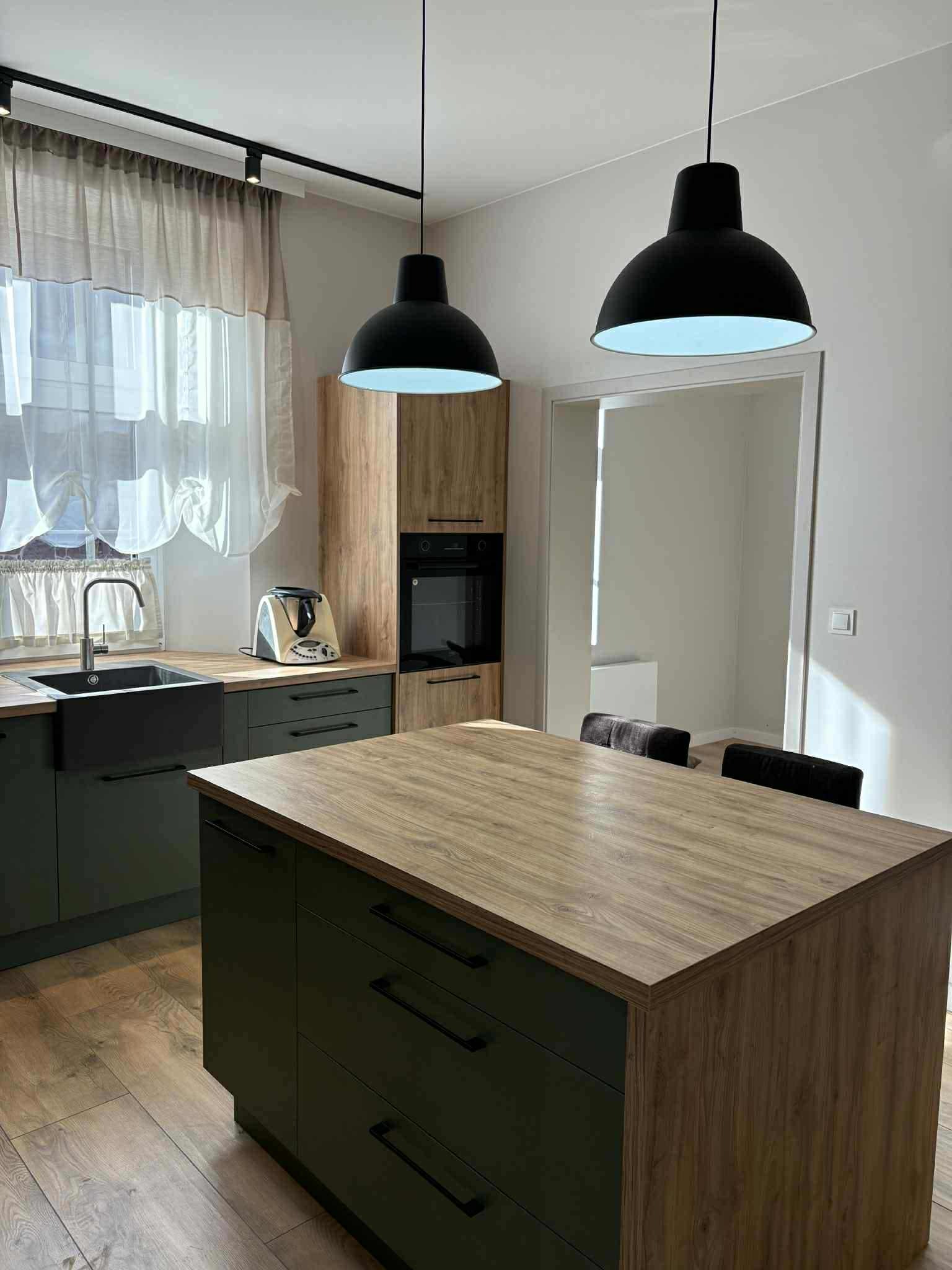 Nowoczesna kuchnia z wyspą w kolorze ciemnej zieleni i drewnianym blatem, oświetlona wiszącymi lampami w kształcie kopuł. Widok na okno i sprzęt AGD.
