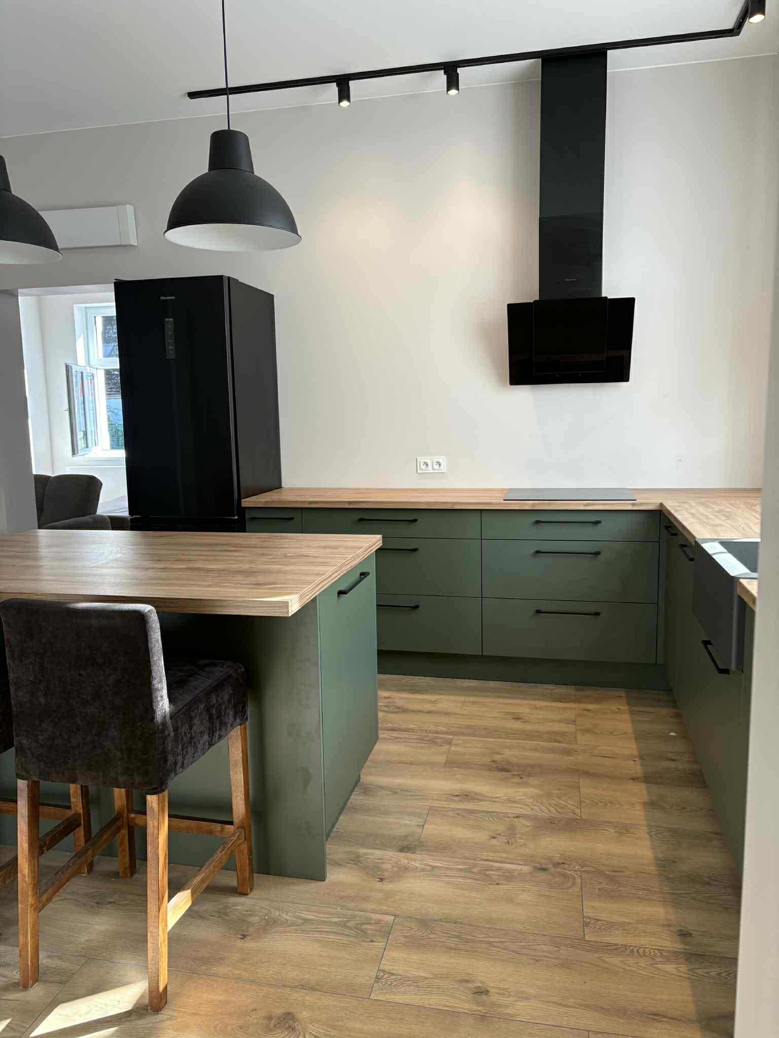 Nowoczesna kuchnia w odcieniach zieleni z drewnianym blatem i czarnymi akcentami, w tym okap i lodówka. Widoczne krzesła barowe przy wyspie.